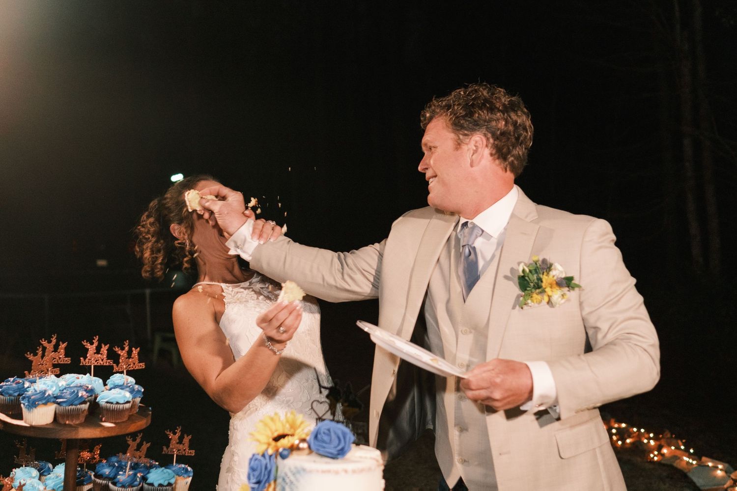 Groom playfully feeds bride wedding cake at elegant outdoor nighttime reception celebration.