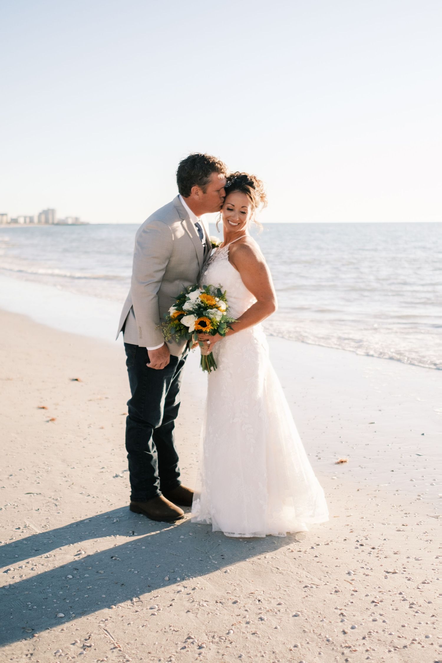 Bride and groom share a romantic kiss on a sunny beach, bride holding a colorful bouquet in a white lace gown.