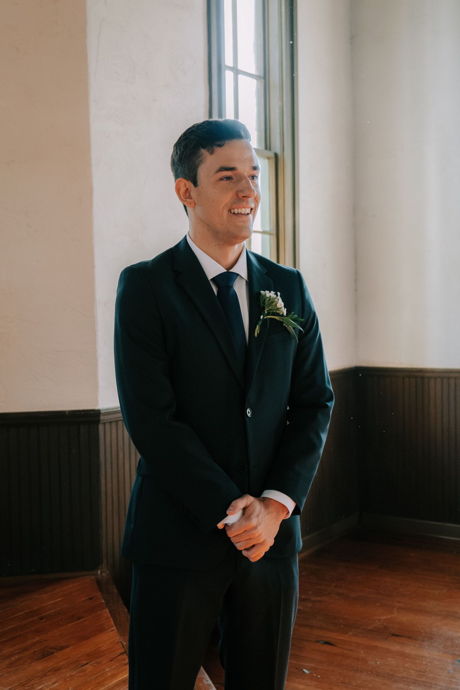 A groom in a classic black suit stands in a room with wooden floors and white walls.