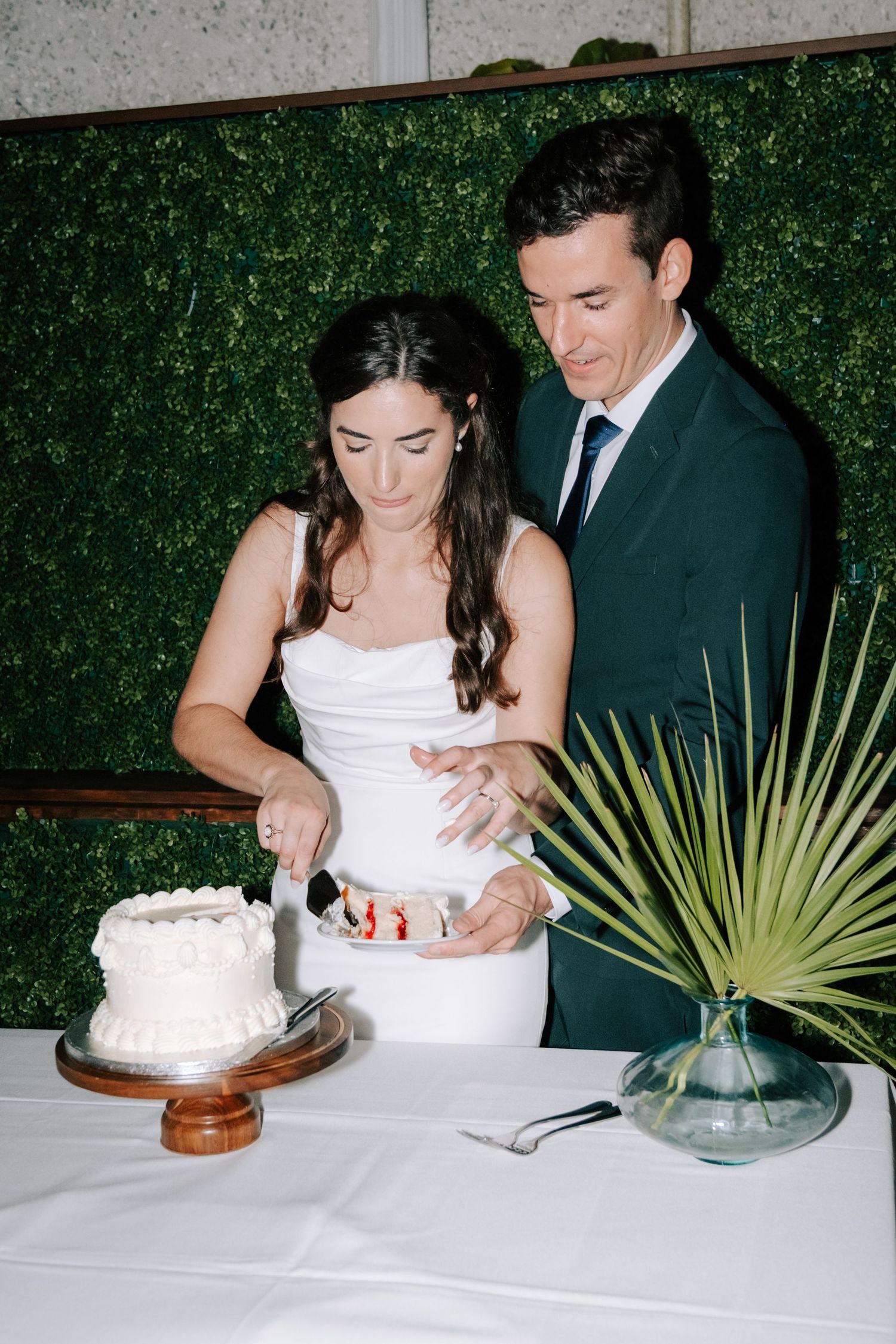 A couple cuts their wedding cake together at a reception with a green hedge wall backdrop.