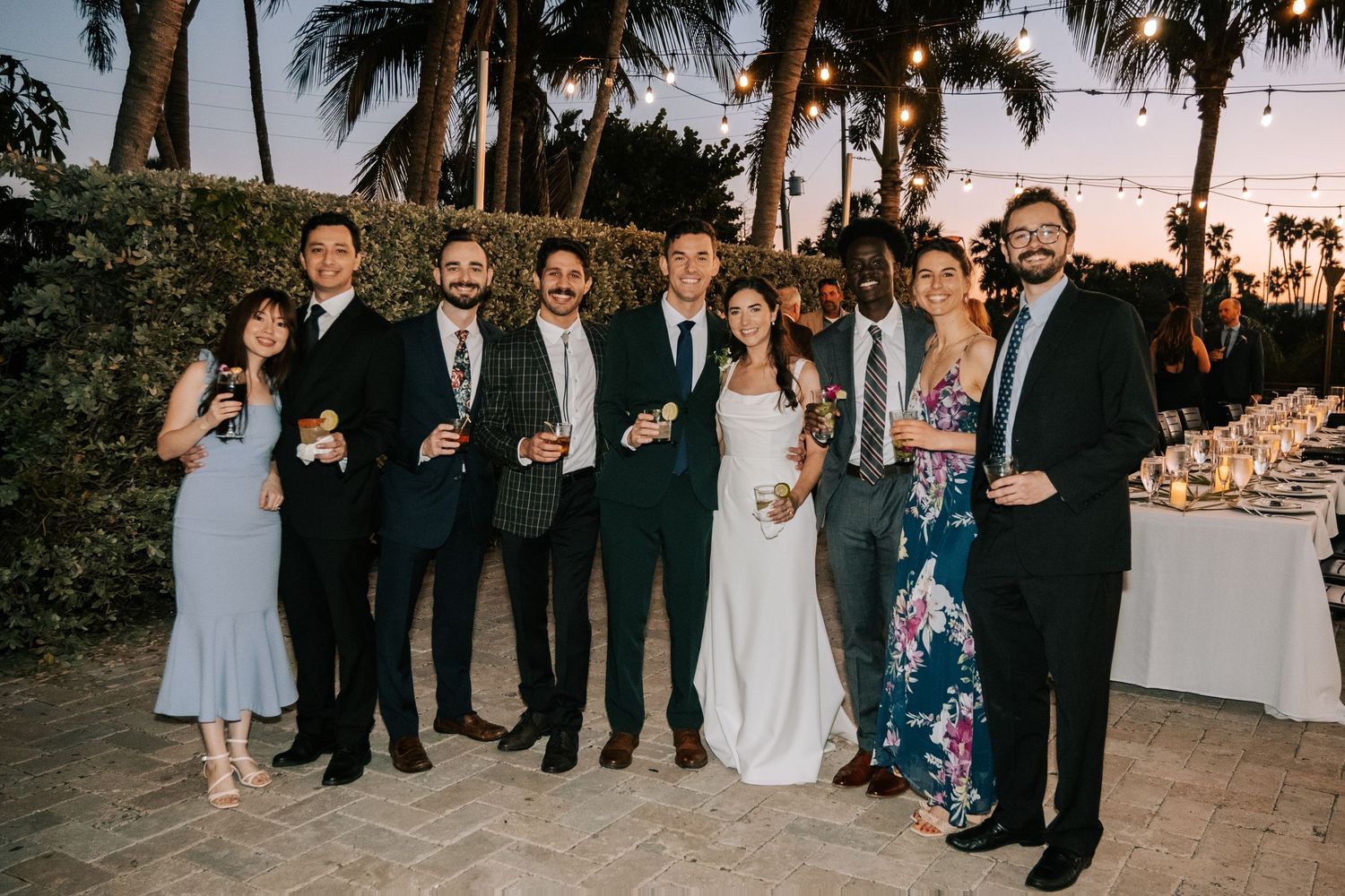 Wedding party members gather for a group photo at sunset with palm trees in the background.