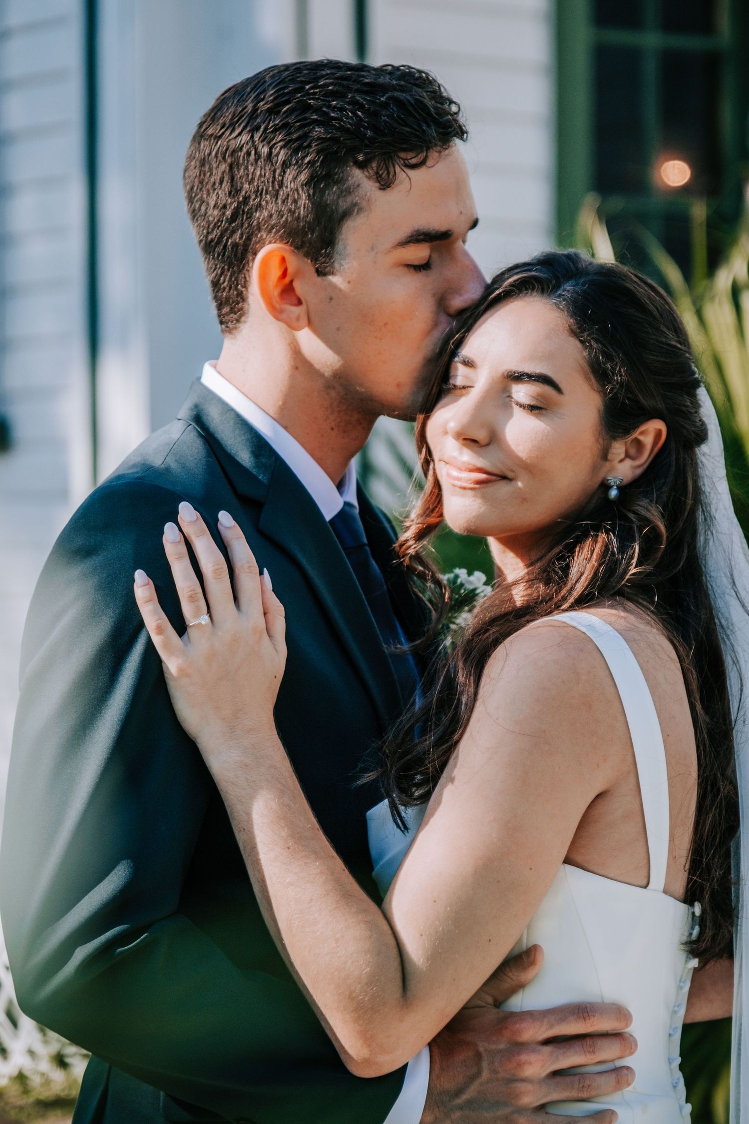An intimate embrace between newlyweds against white church walls in soft sunlight.