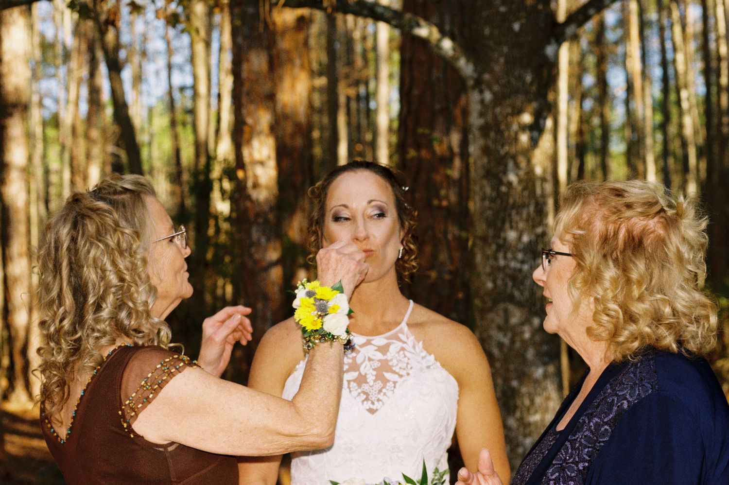 Bride in lace halter gown being touched up by two women in pine forest setting.