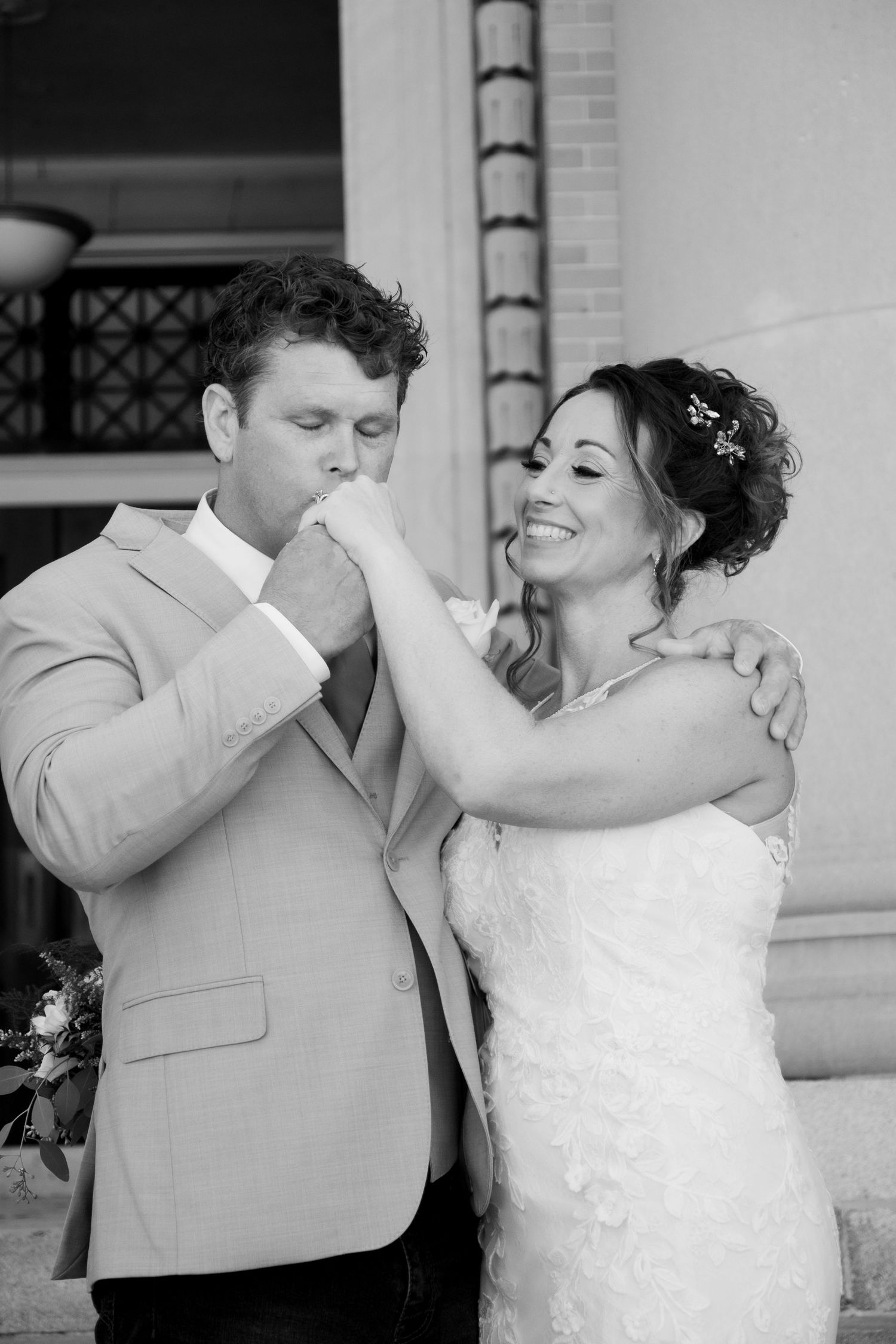 Groom kisses bride's hand as she smiles in a lace wedding dress, black and white photo.