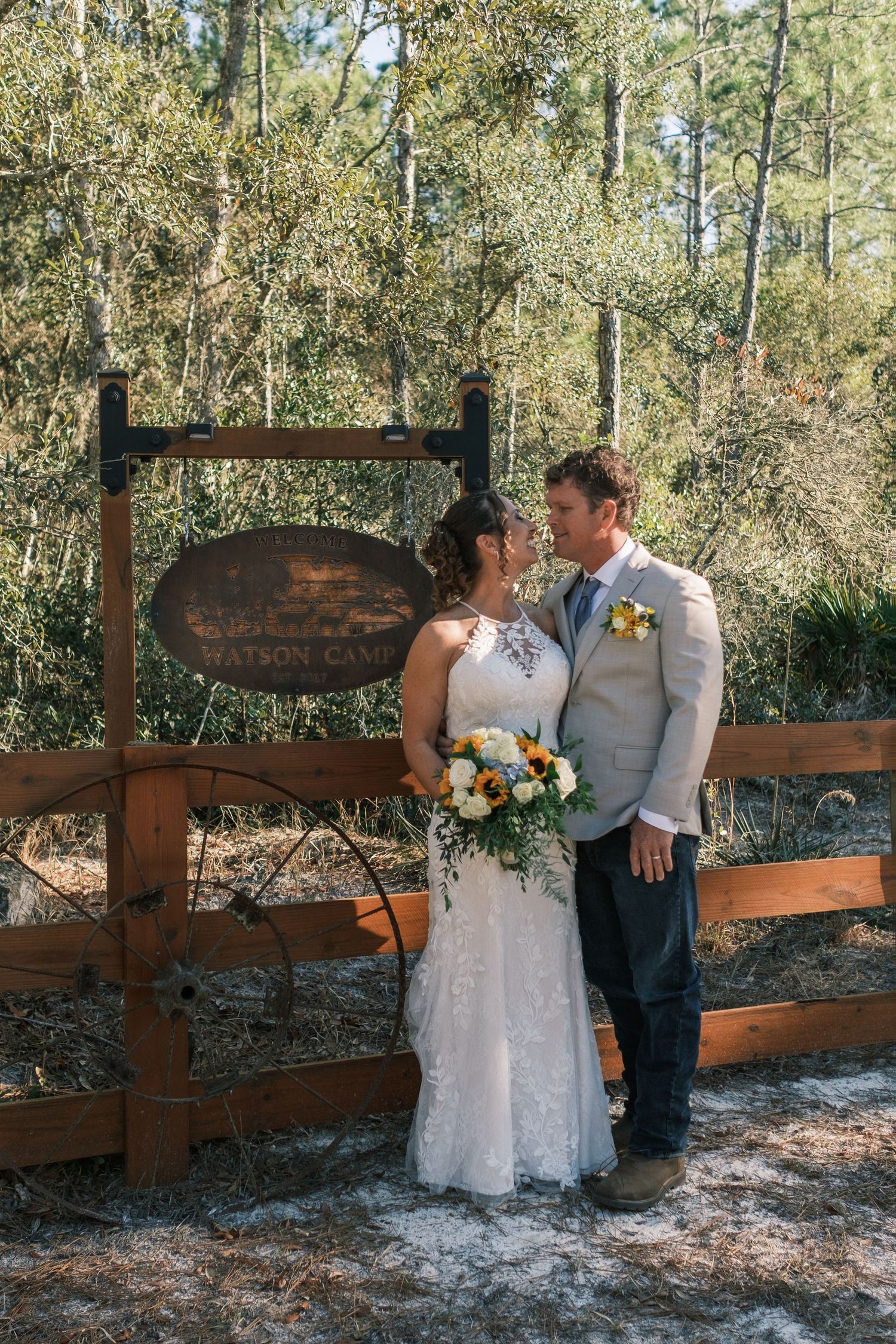 Bride and groom share a tender kiss in front of a rustic wooden Watson Camp sign surrounded by trees.