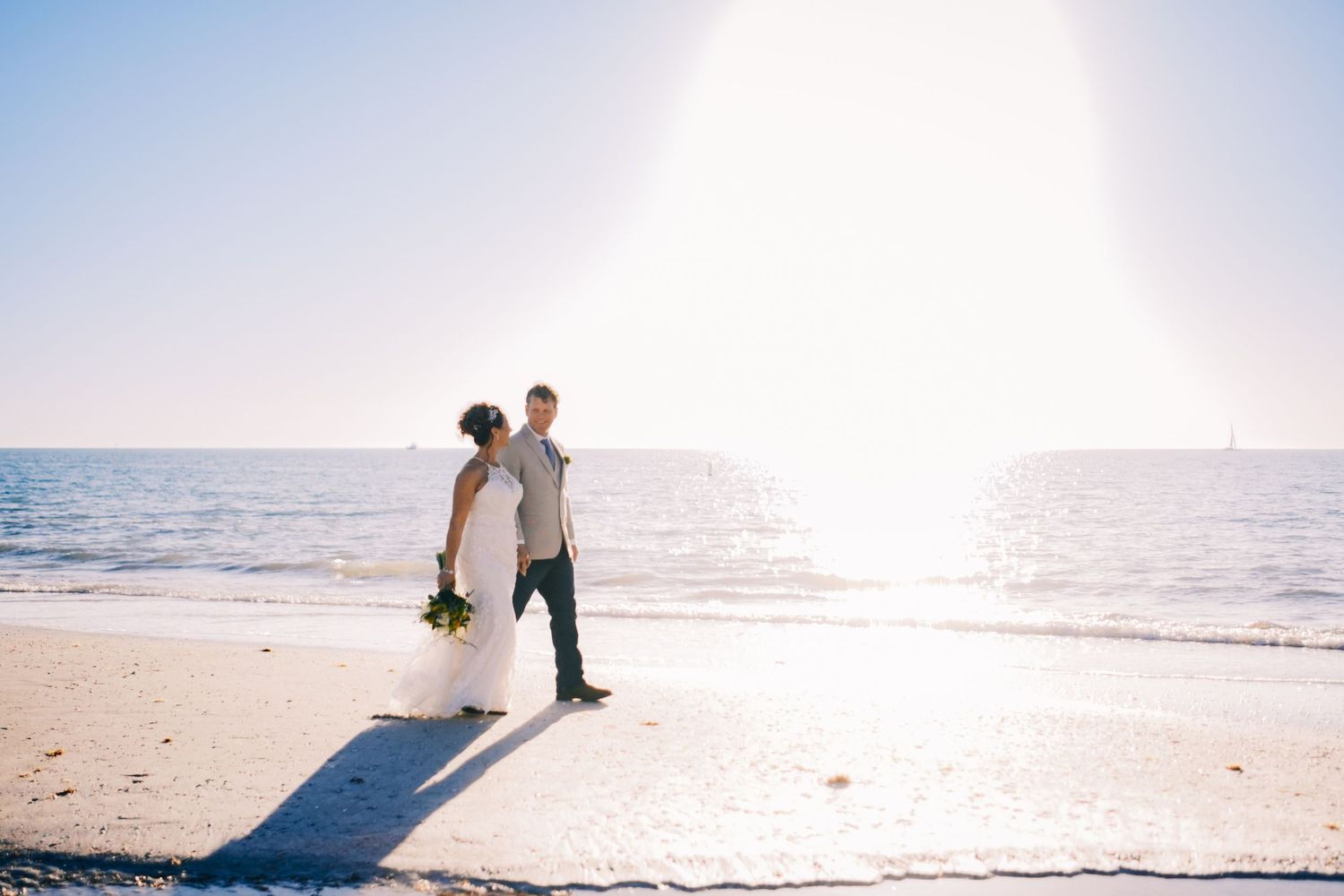 Bride and groom stand back to back on a sunlit sandy beach with sparkling ocean behind them.