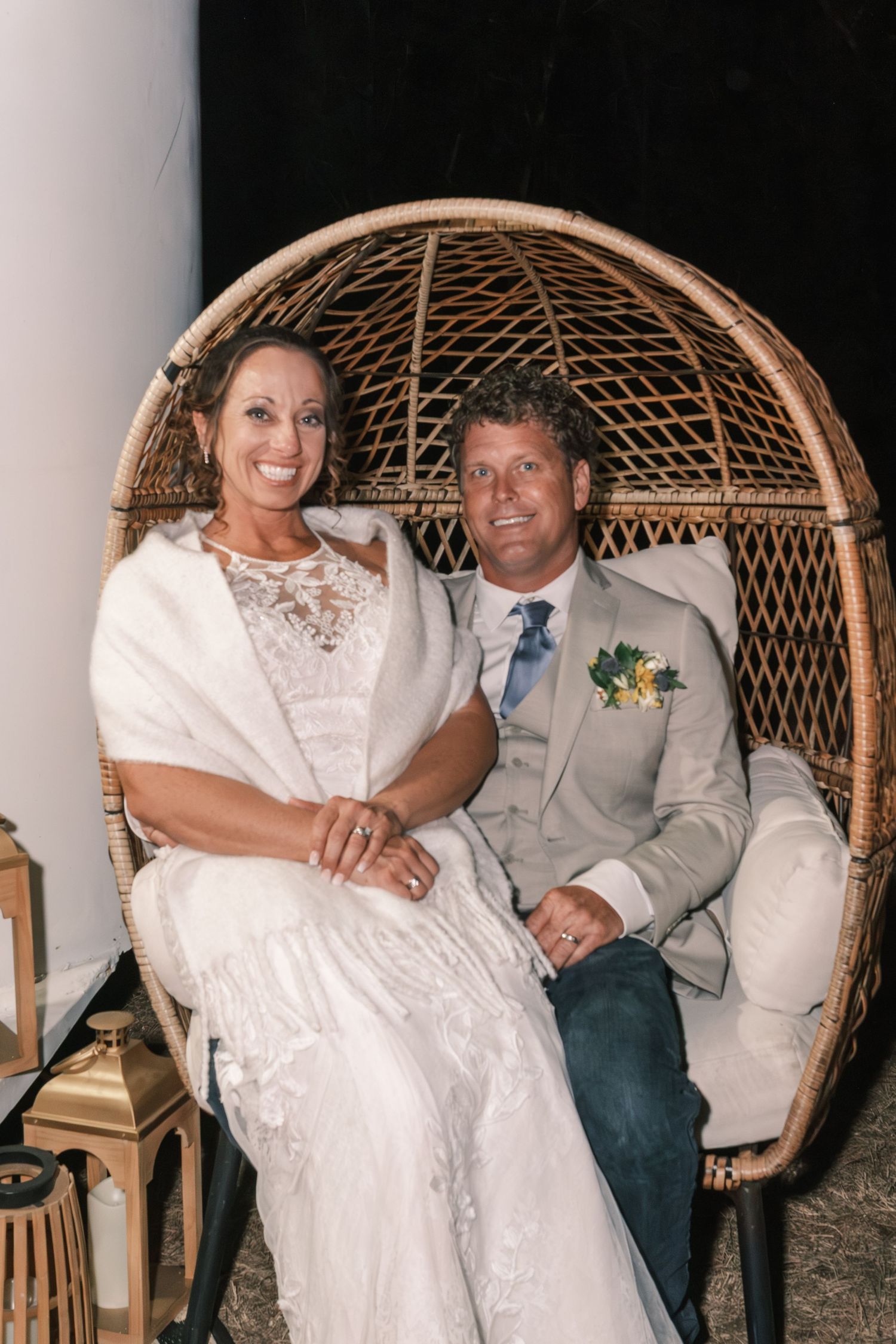 Bride and groom sitting together in wicker egg chair at wedding reception smiling at camera.