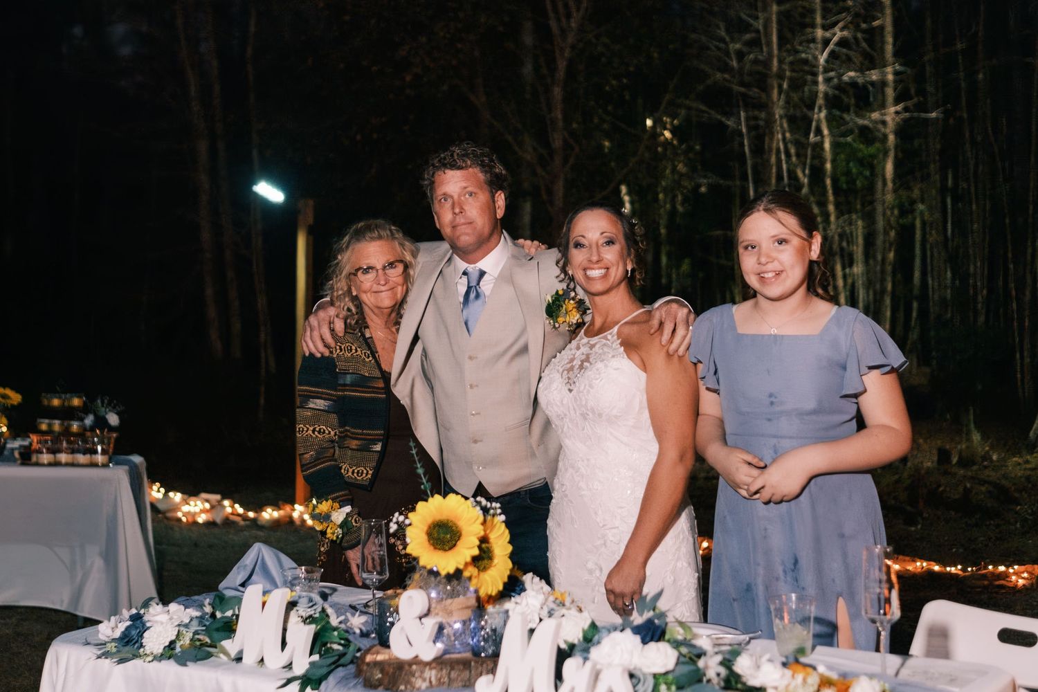 Newlyweds pose with family members at a decorated wedding reception table with sunflowers.