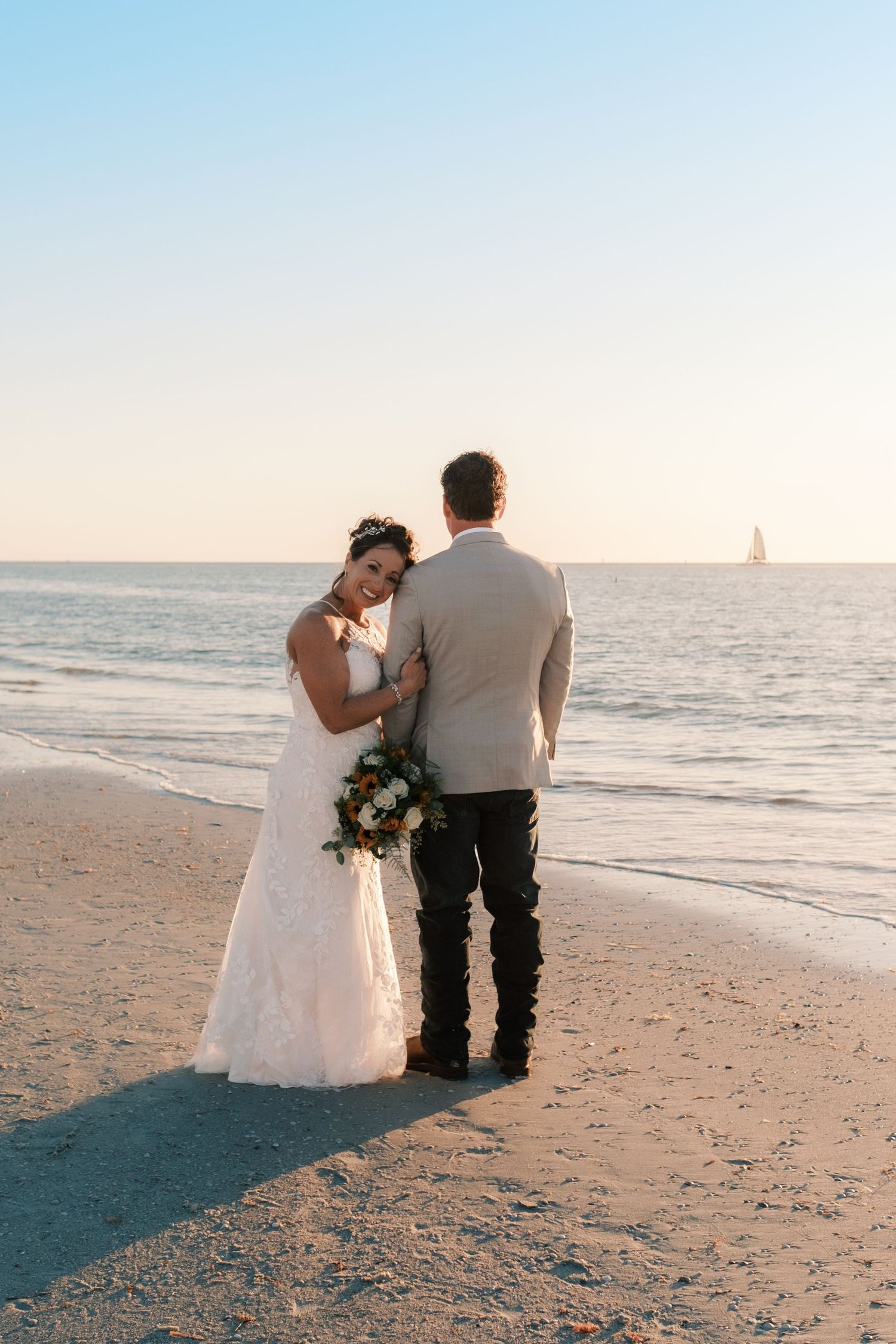 Bride and groom stand together on beach at sunset with sailboat visible on the horizon.