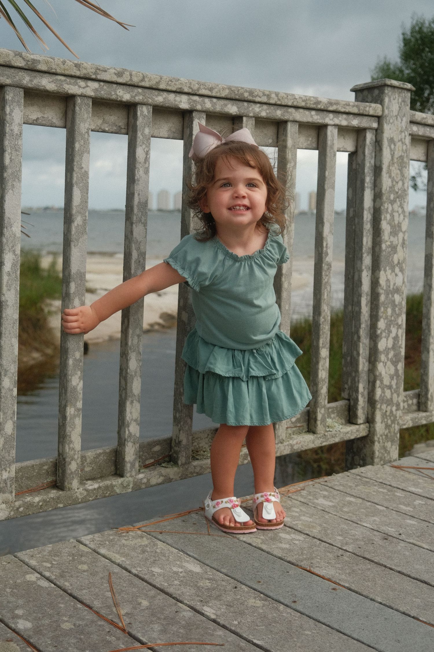 A small child wearing a green ruffled dress plays on a weathered wooden deck overlooking water.