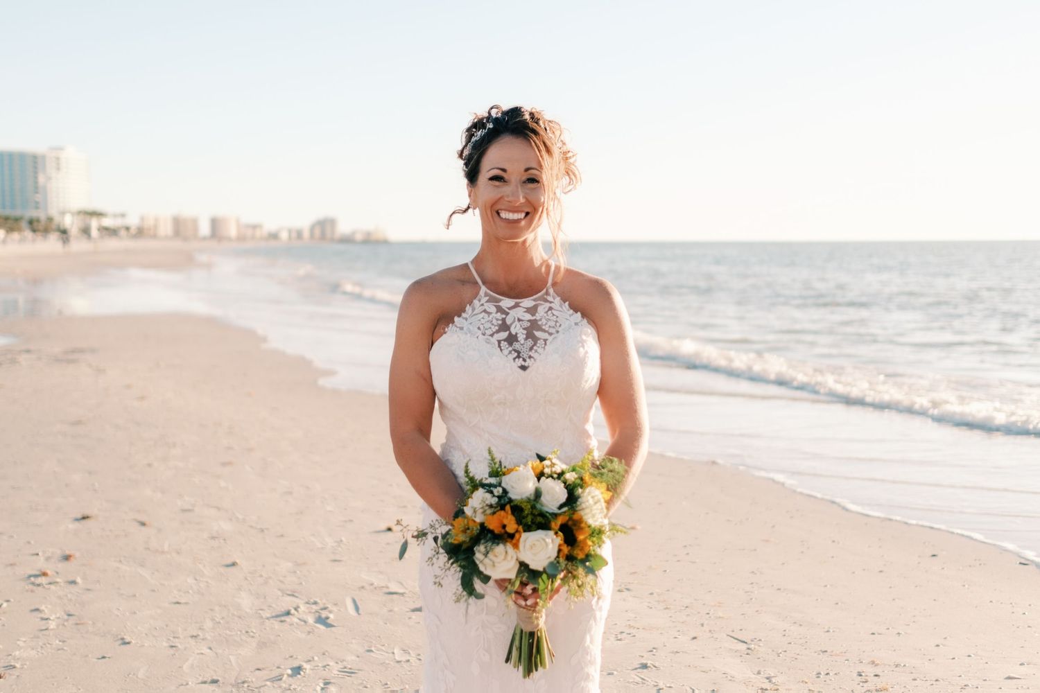 Radiant bride smiles on a sunny beach holding a colorful bouquet of white and yellow flowers.