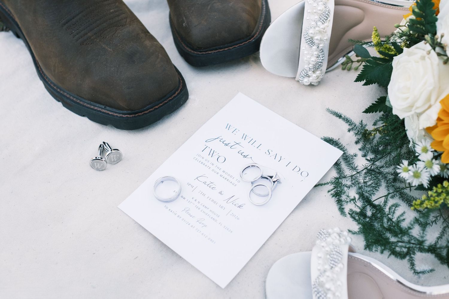 Wedding flat lay featuring invitation, rings, cowboy boots, and colorful bridal bouquet.