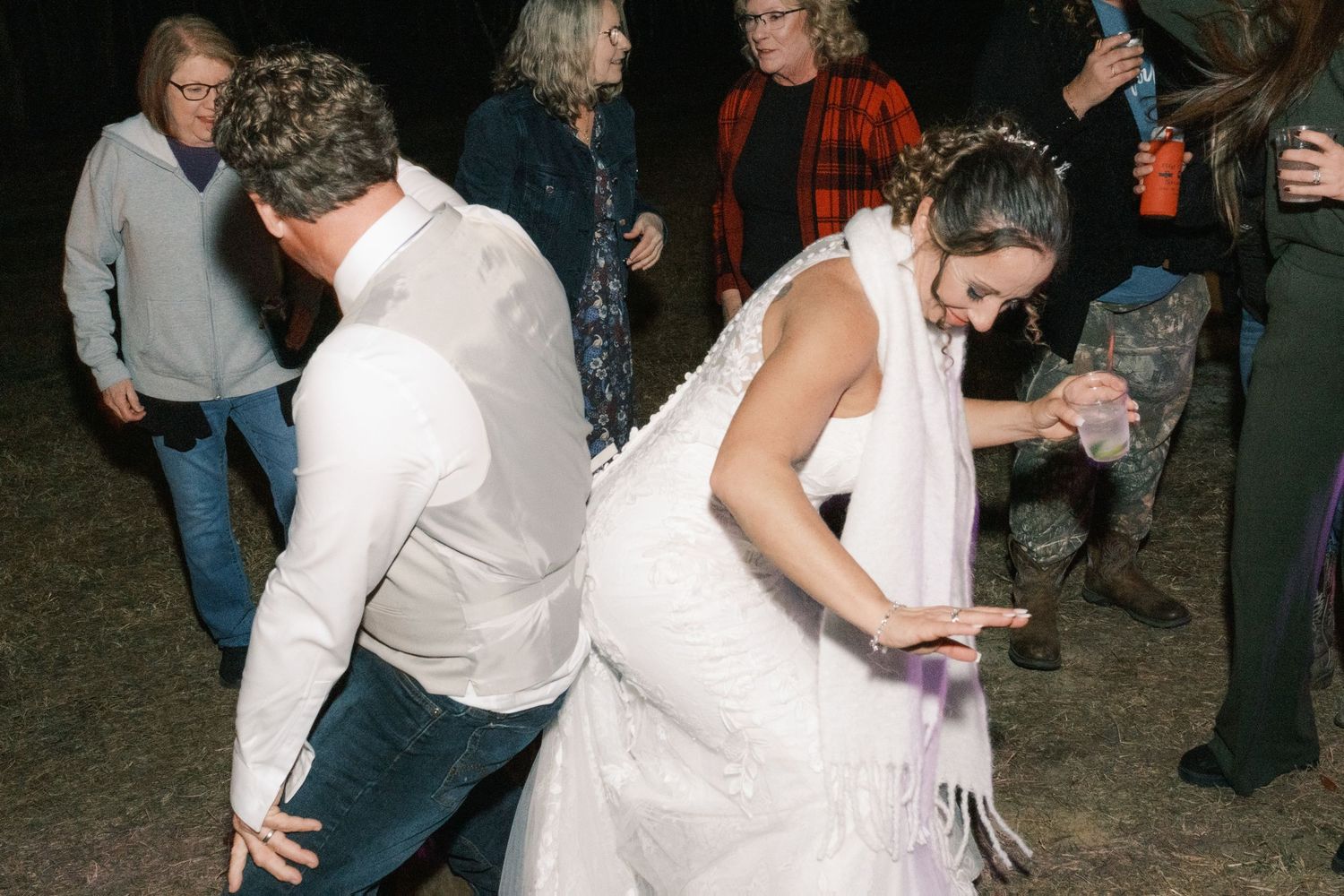 Bride in white gown and groom dance energetically with wedding guests at outdoor night reception.
