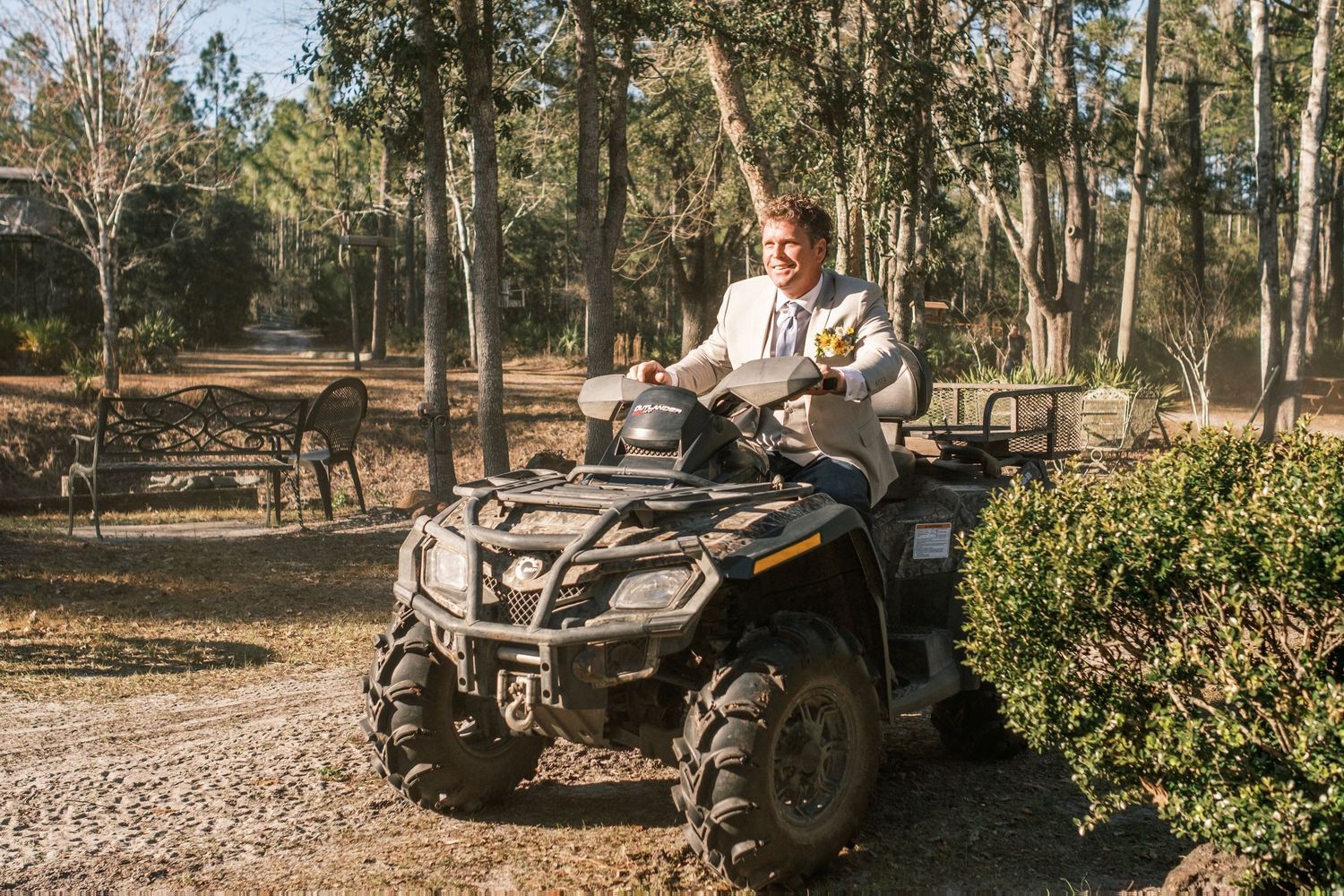 Man in white shirt rides black ATV quad bike through wooded rural property on sunny day.