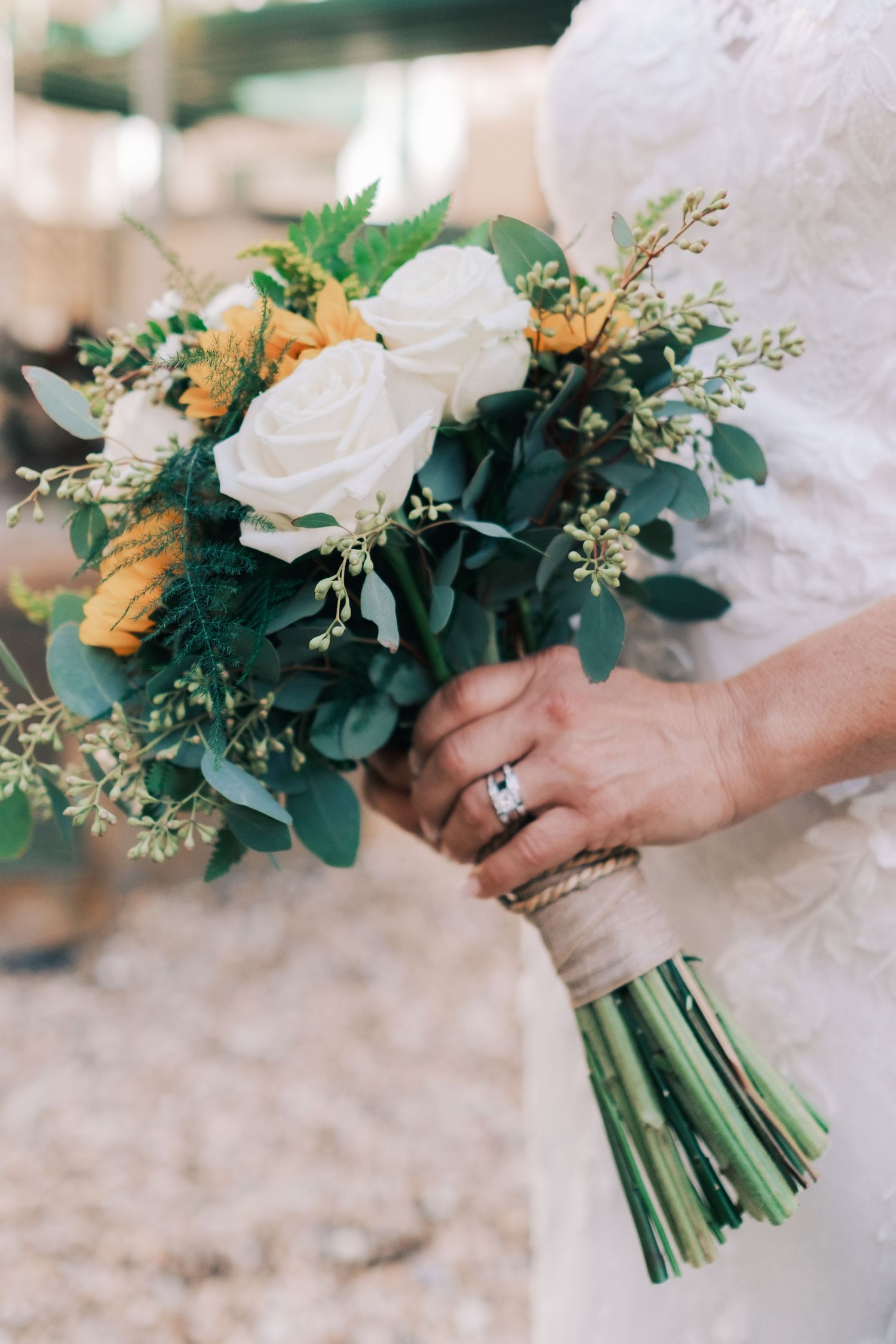 Bride holds elegant bouquet of white and yellow roses with greenery, wearing diamond ring.