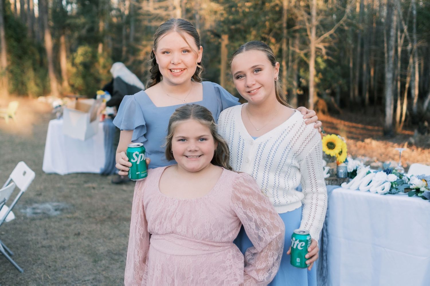Three young girls in blue and pink dresses smiling at an outdoor wedding reception holding Sprite cans.