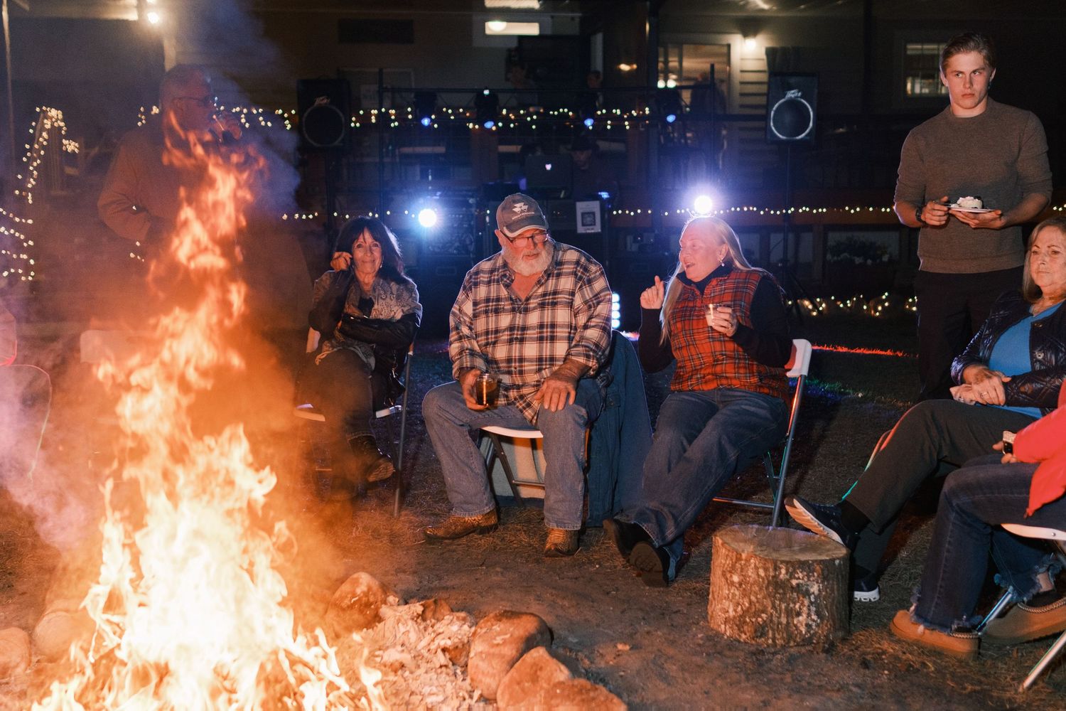 Wedding guests gathered around a large crackling bonfire at an outdoor nighttime wedding reception.