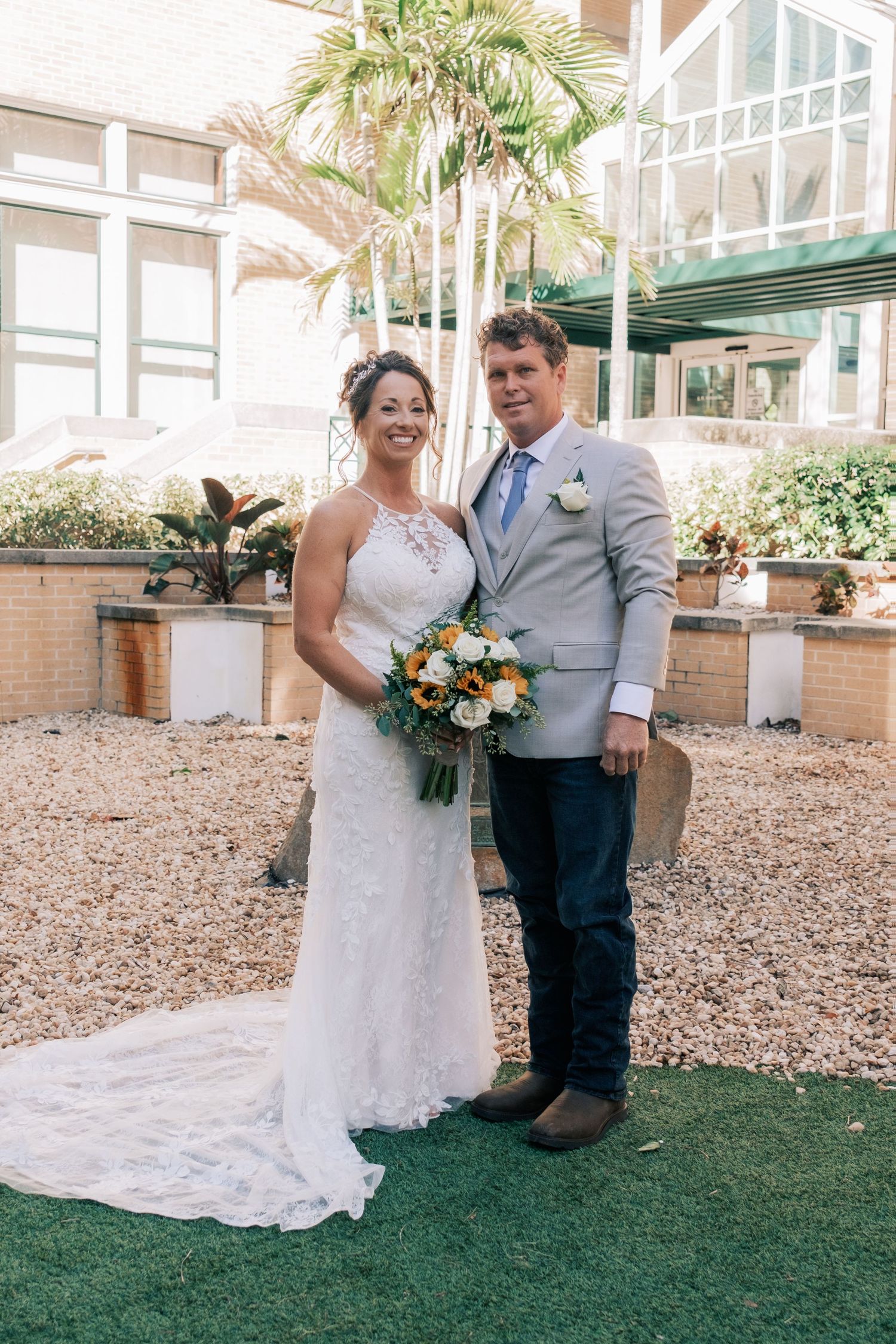 Smiling bride and groom pose together in a courtyard, bride holding a white and orange bouquet.