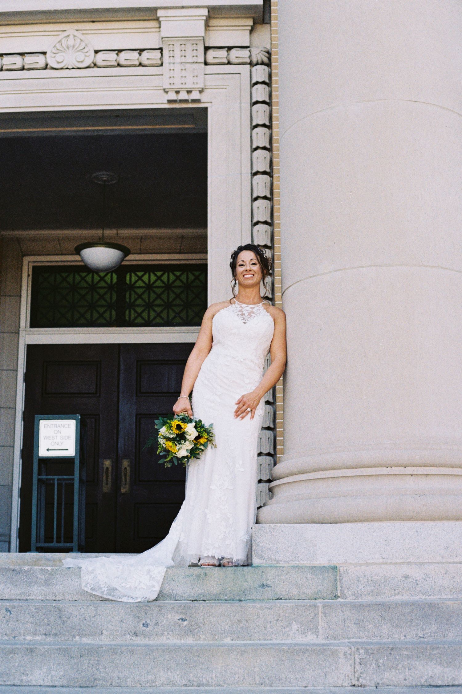 Bride in lace halter gown holding sunflower bouquet posing on steps of historic building.