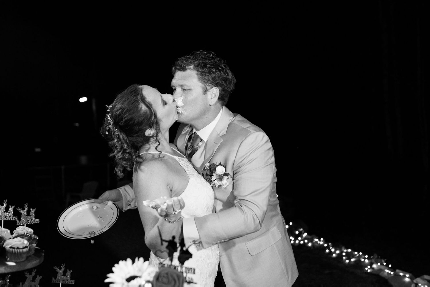 Black and white photo of a bride and groom sharing a kiss while holding wedding cake plates at their reception.