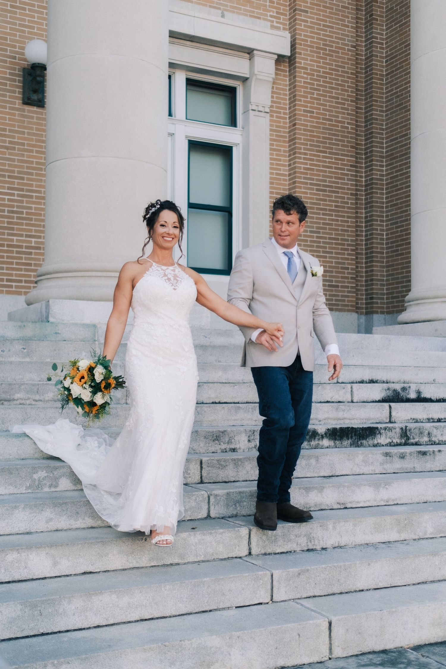 Bride and groom hold hands walking down stone steps outside a brick building on their wedding day.