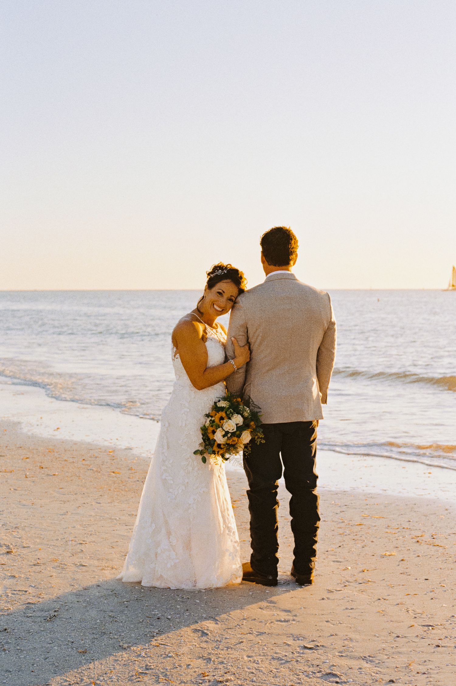 A bride in a white lace gown embraces her groom while standing on a sandy beach at sunset.