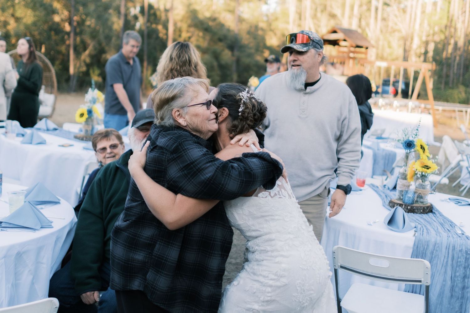Bride in lace gown shares a warm embrace with a guest at an outdoor wedding reception.