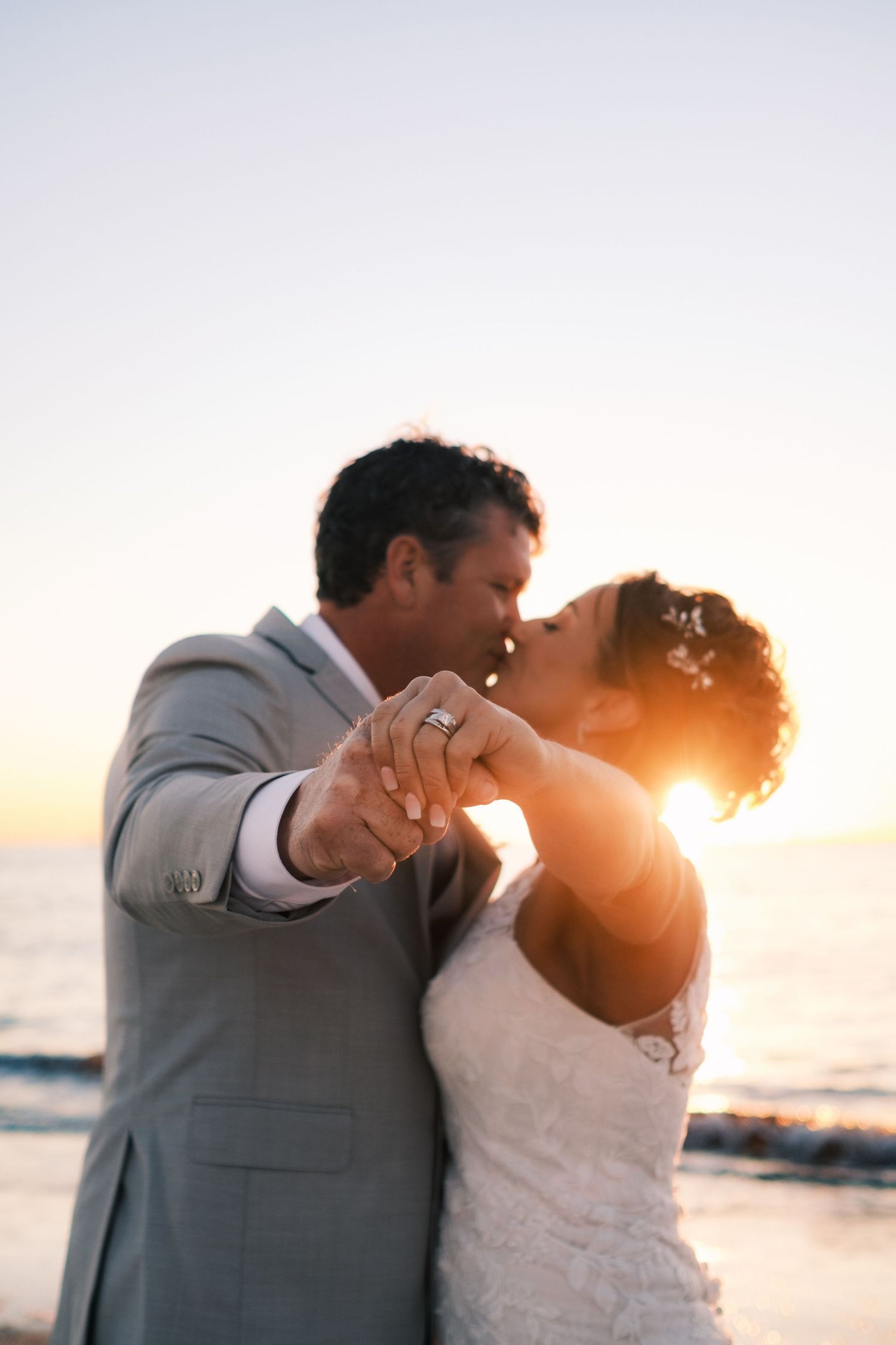 Bride and groom share a romantic sunset kiss on the beach, displaying wedding rings with golden light behind them.