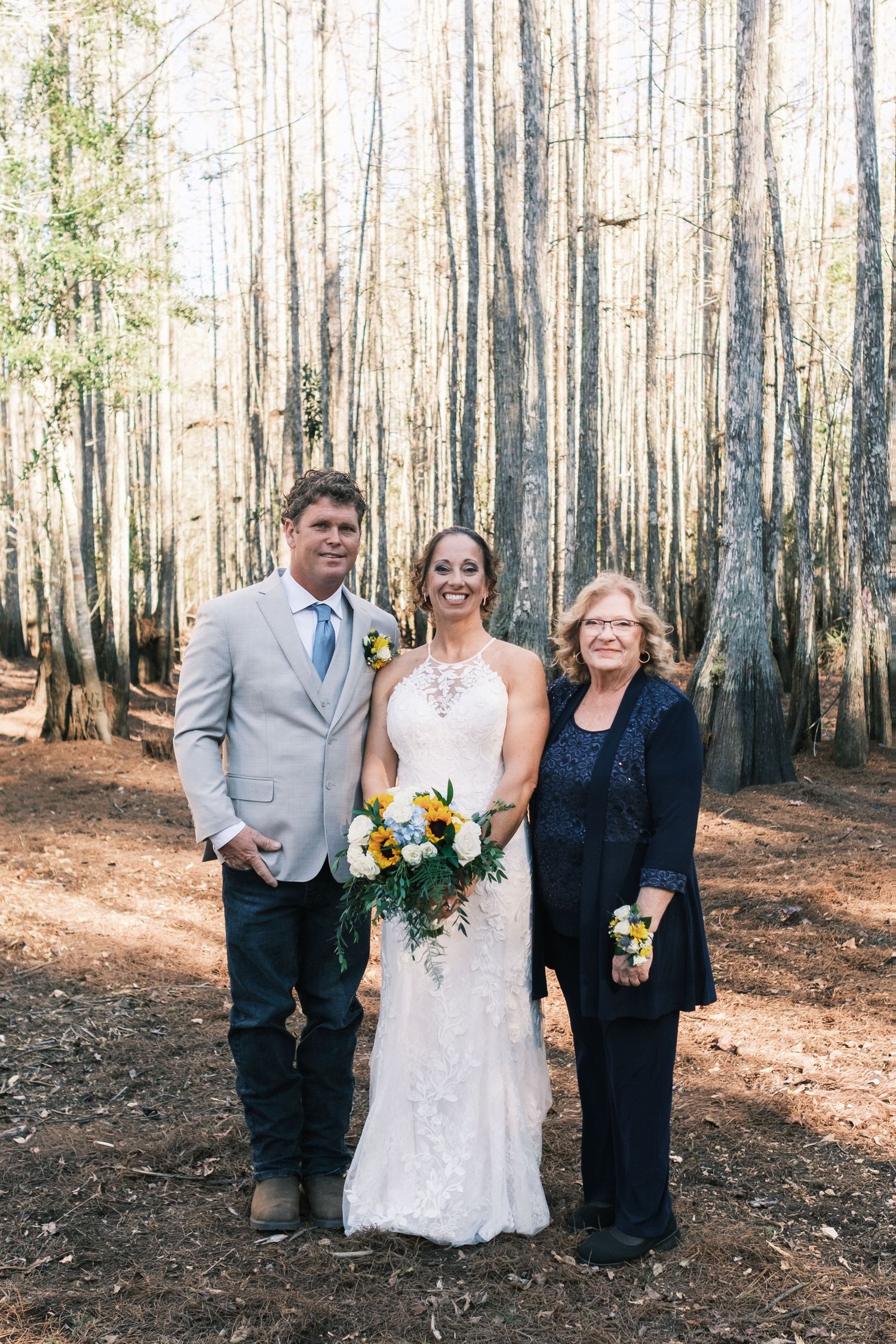 Bride in white lace gown with bouquet poses with two guests in a sunlit forest of tall slender trees.