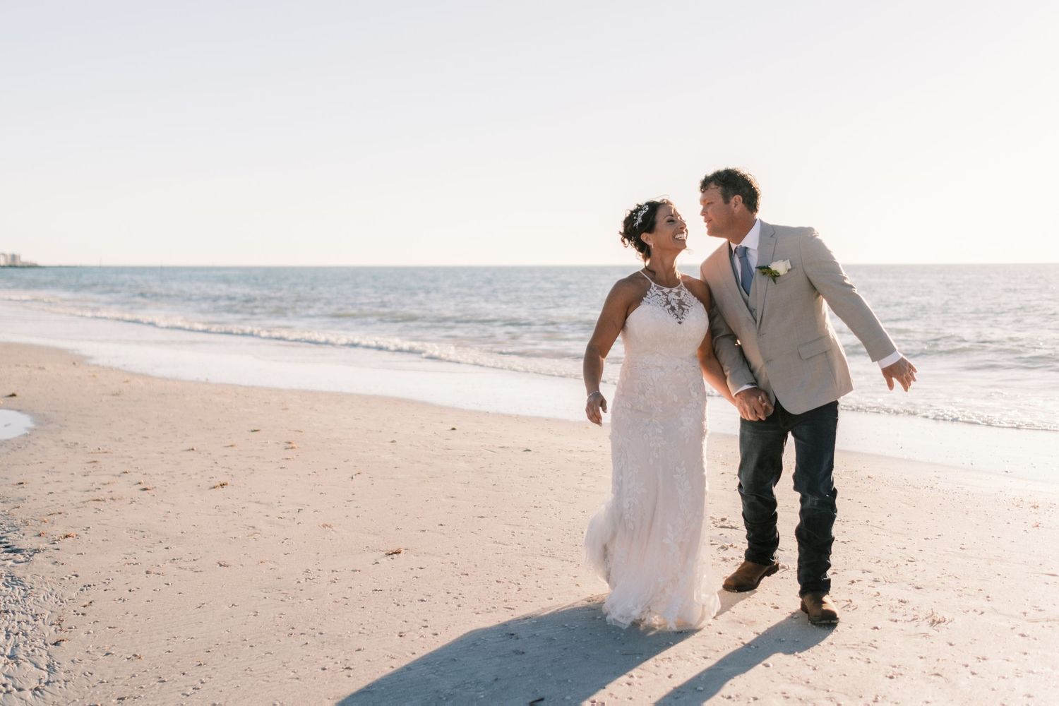Bride and groom laugh together walking on sandy beach at sunset in wedding attire.
