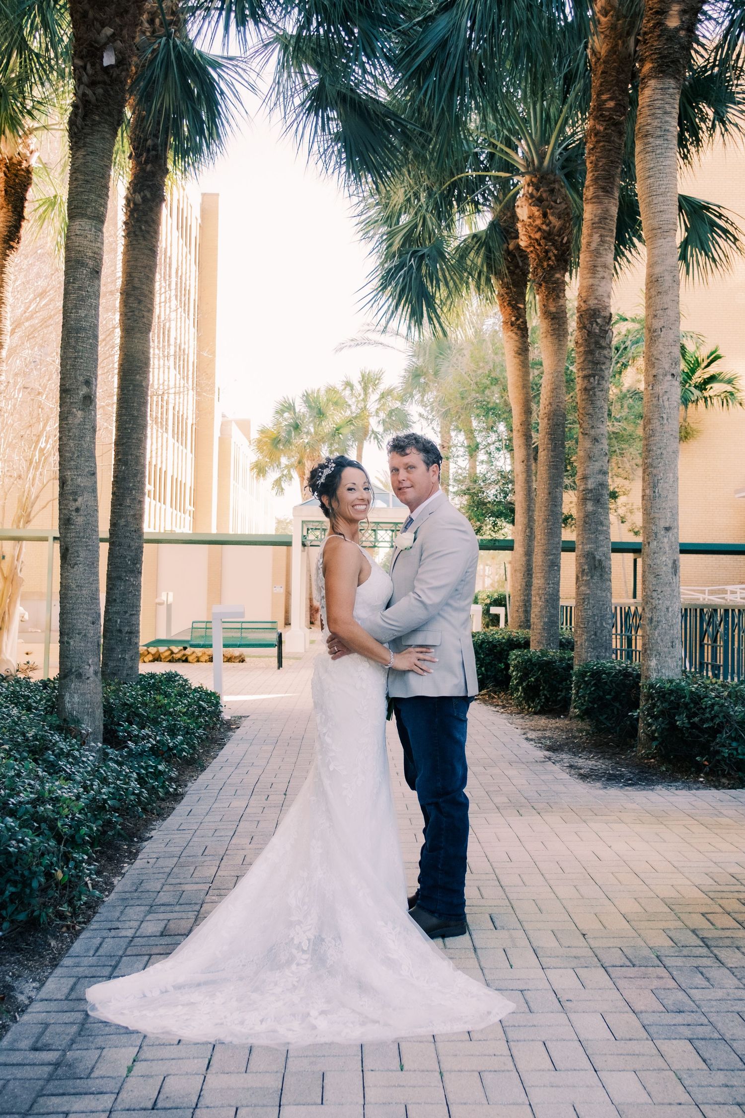 Bride and groom embracing among tropical palm trees in elegant wedding attire outdoors.