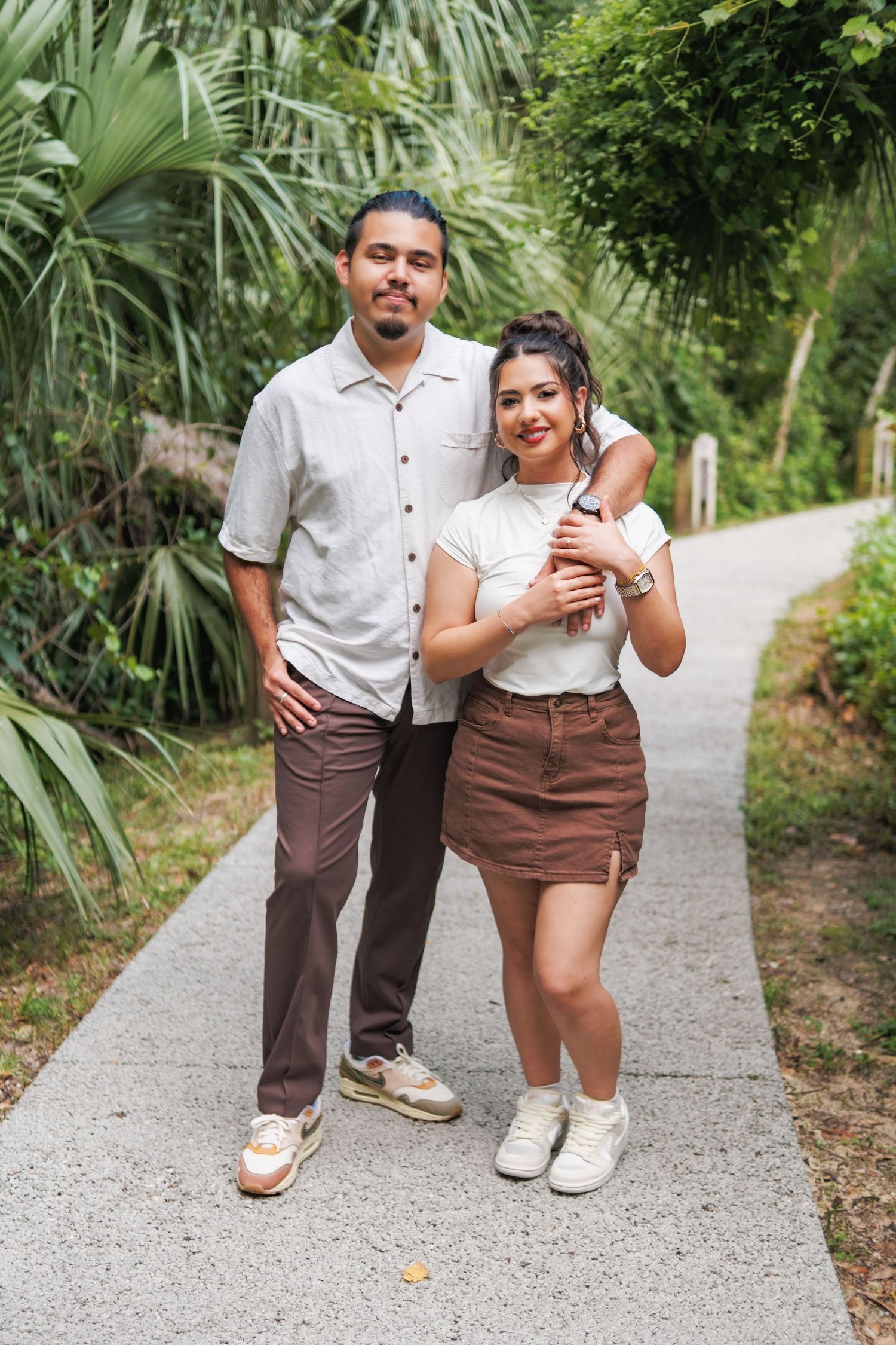 A couple in coordinating brown and white outfits walks along a winding garden path surrounded by palm trees.
