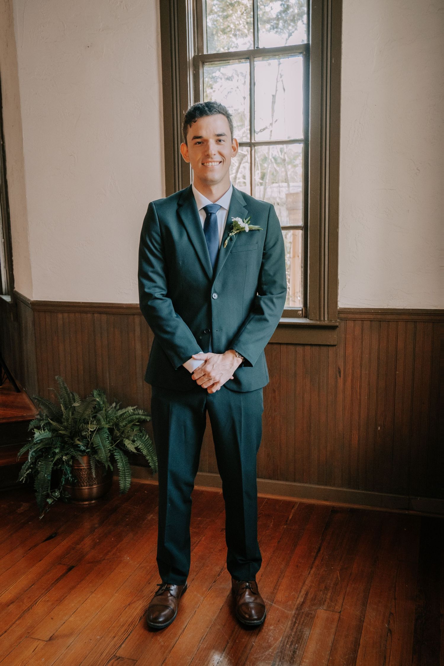 A groom in a navy suit poses on hardwood floors in a room with wood paneling and windows.