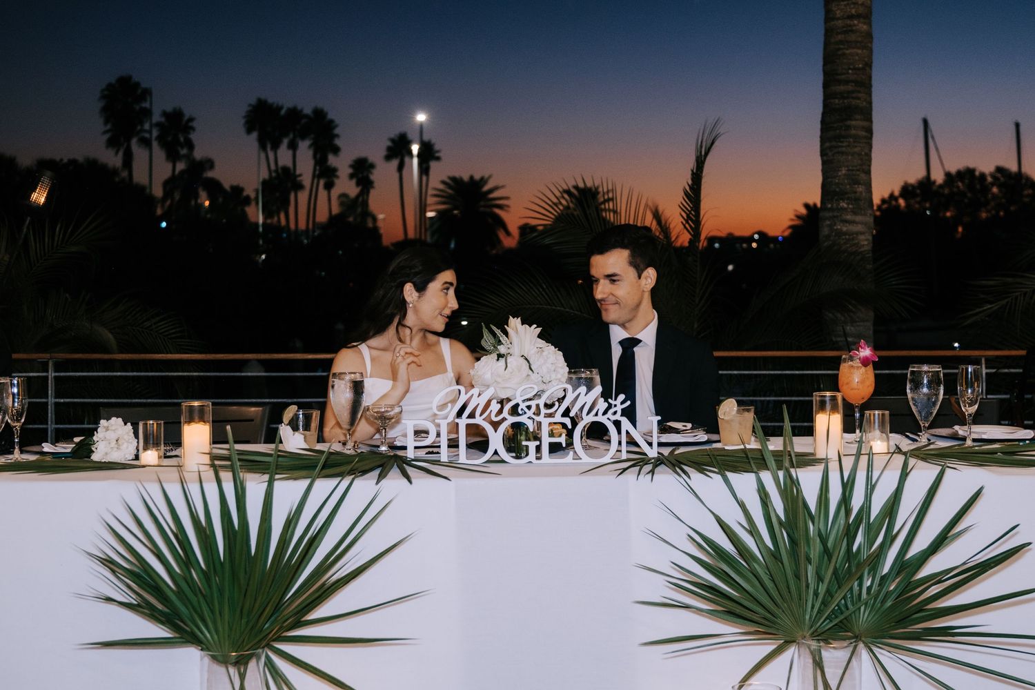 A twilight wedding reception setup with palm fronds and decorative lighting outdoors.