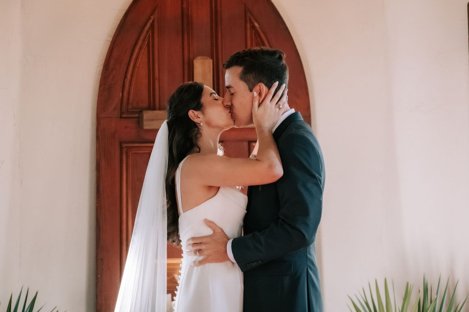 A couple shares a romantic kiss in front of white curtains and wooden doors during a wedding ceremony.
