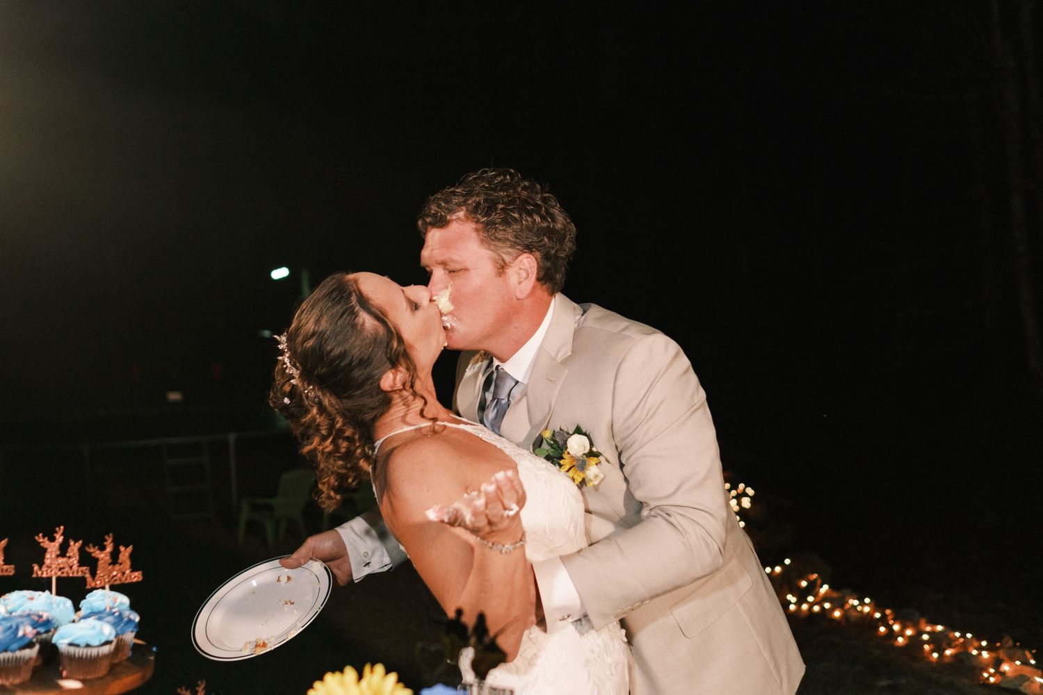 Bride and groom sharing a romantic dip kiss at their outdoor nighttime wedding reception near a dessert table.