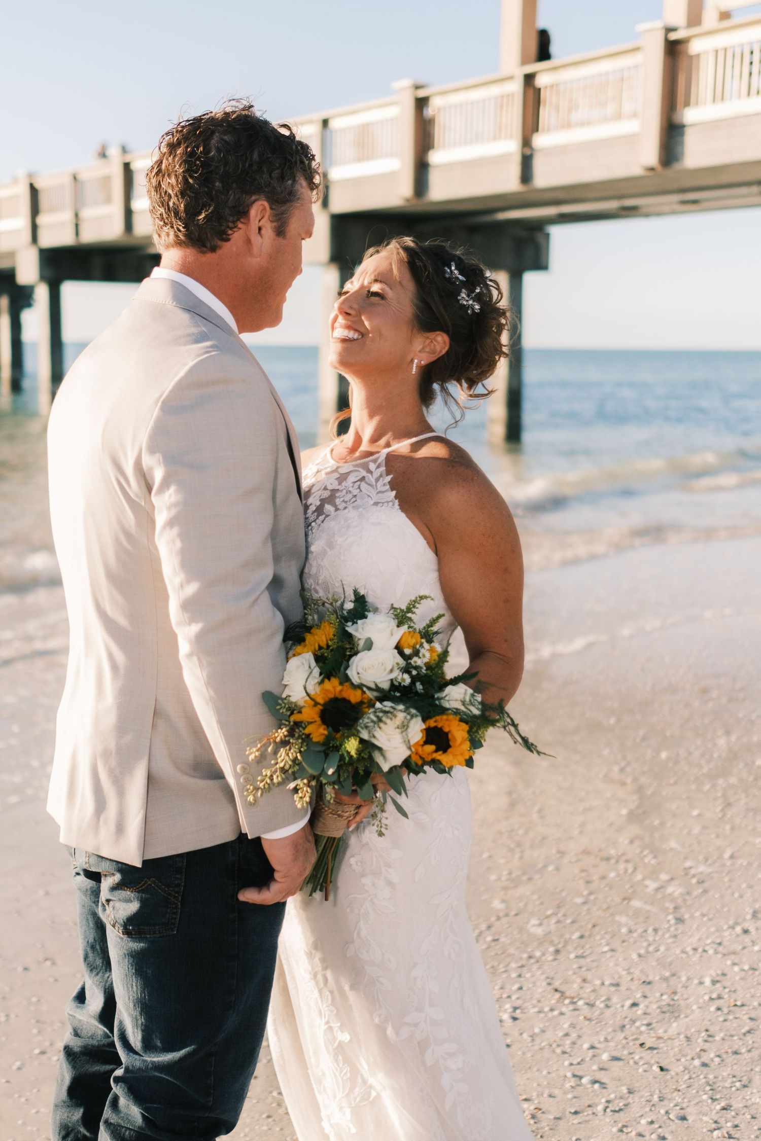 Bride and groom embrace on a sunny beach near a pier, bride holding a sunflower and white rose bouquet.