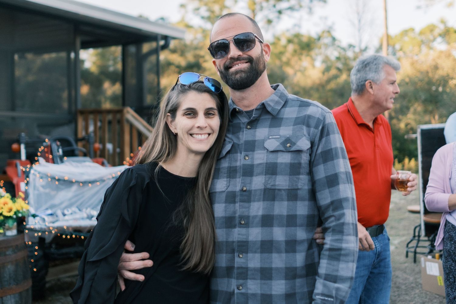 Smiling couple poses together at an outdoor wedding event, man in plaid shirt with arm around woman in black.