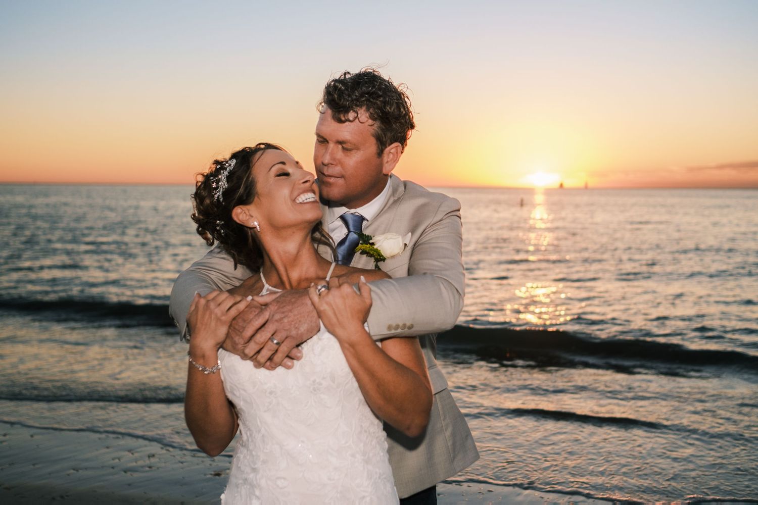 Groom embraces bride from behind at sunset on the beach, both laughing with ocean waves in background.