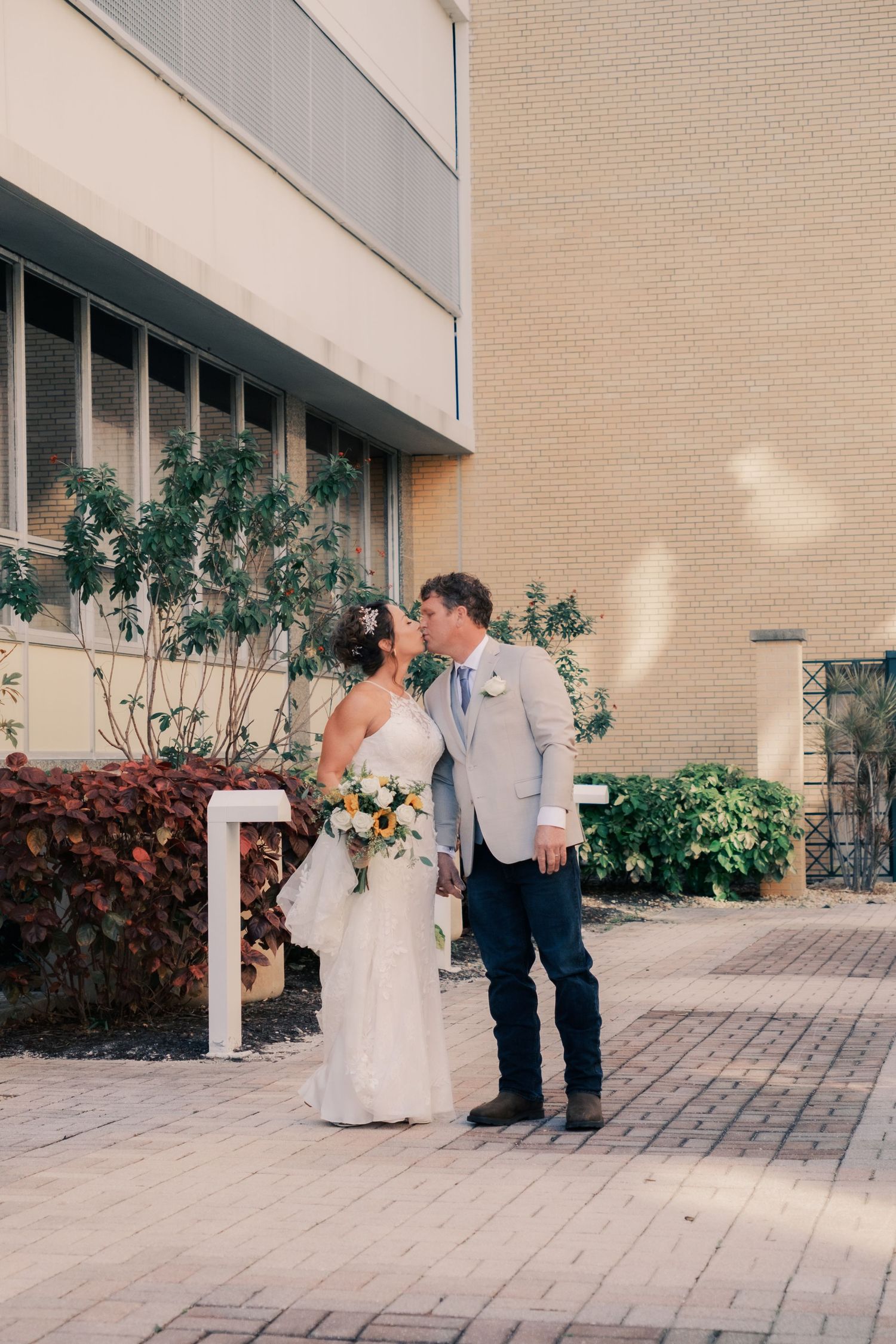Bride and groom share a kiss outside a brick building on their wedding day, bride holding a yellow and white bouquet.