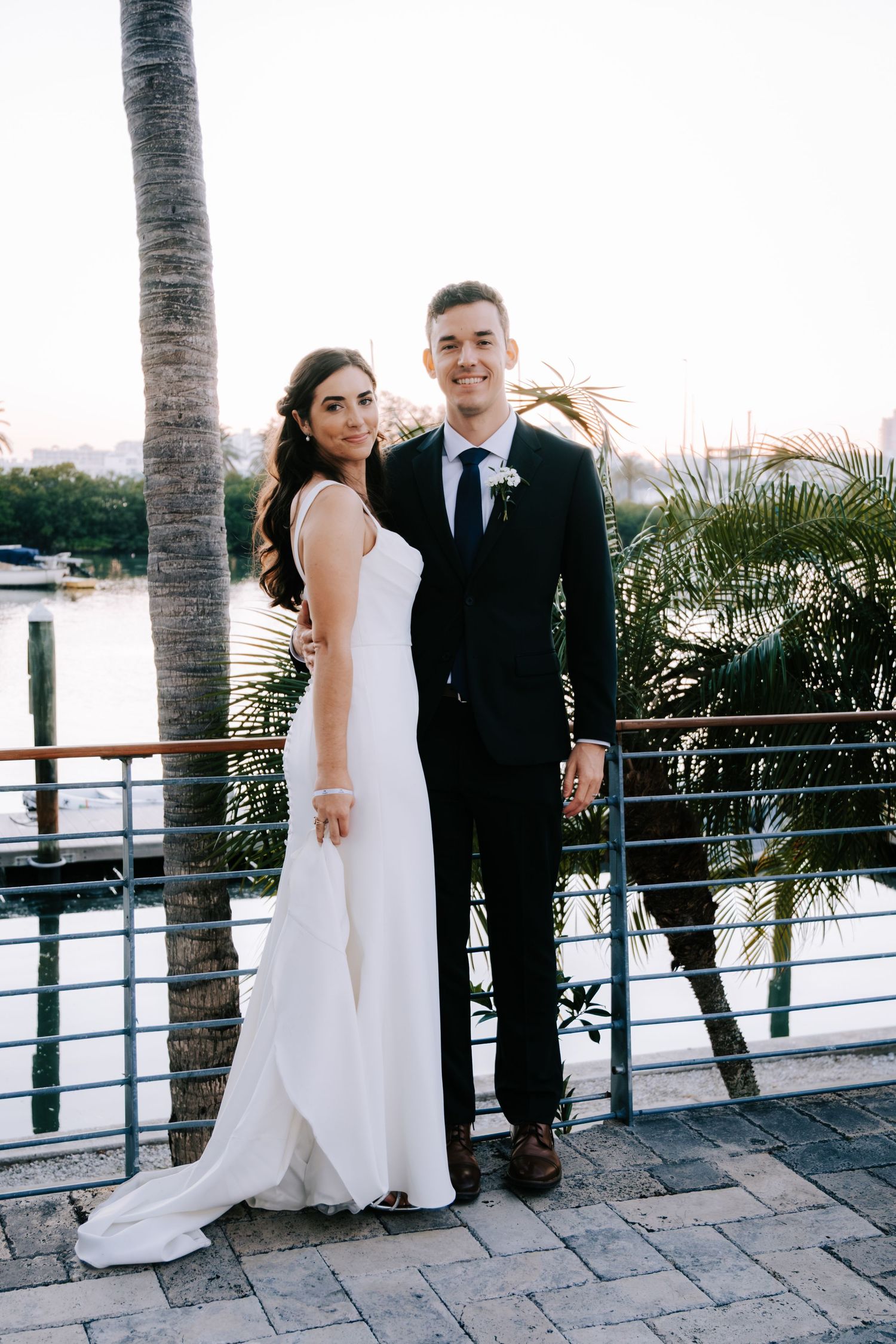 A waterfront wedding photoshoot with palm trees and a railing in the background at sunset.