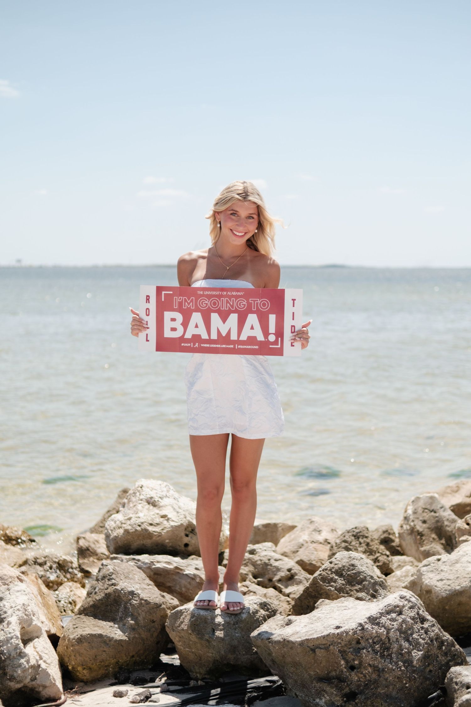 Person in white dress standing on coastal rocks holding a red Bama sign with ocean in background.