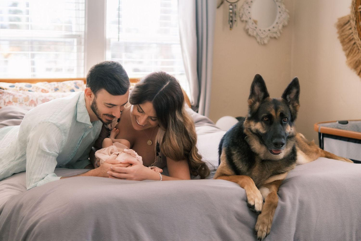 A couple lies on a bed with their newborn baby while a German Shepherd dog sits beside them in a cozy bedroom.