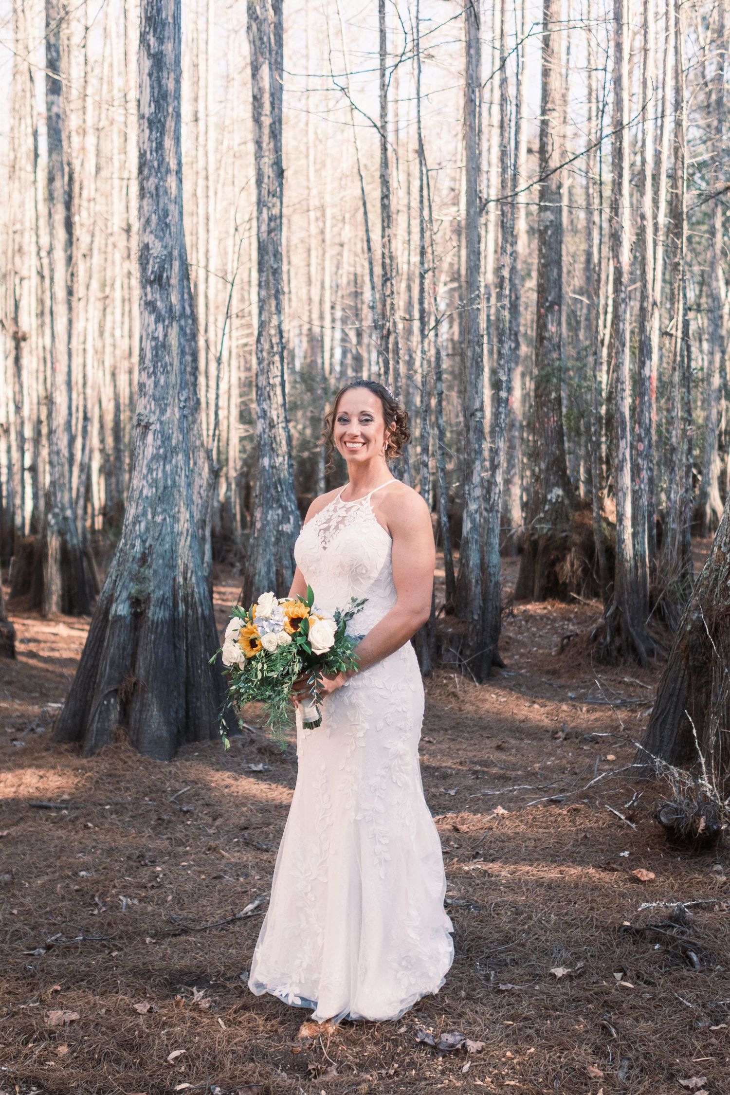Bride in lace mermaid wedding gown holding bouquet of white and orange flowers in a forest setting.