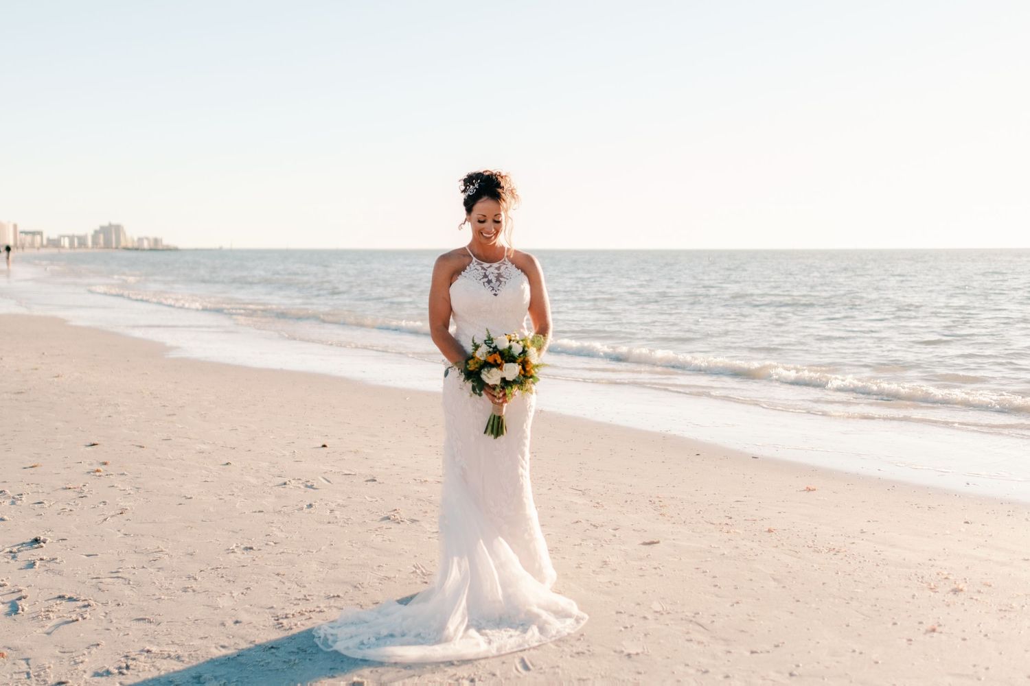 Bride in halter wedding gown holds colorful bouquet while standing on a serene beach.