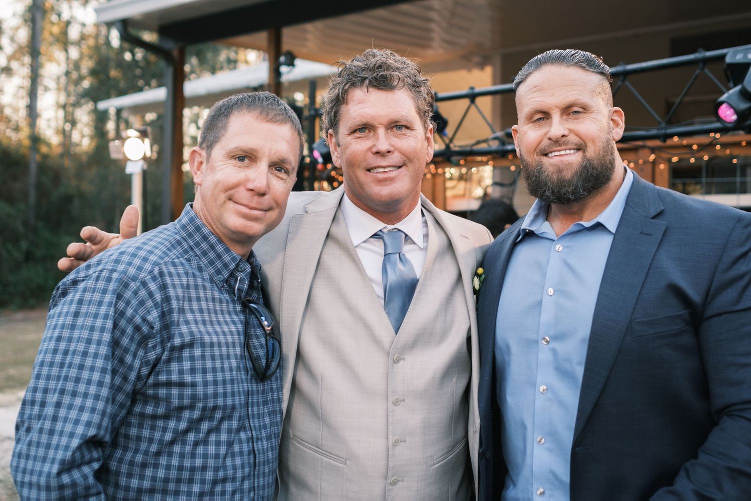 Groom in a tan suit smiles warmly while posing with two friends at an outdoor wedding reception.