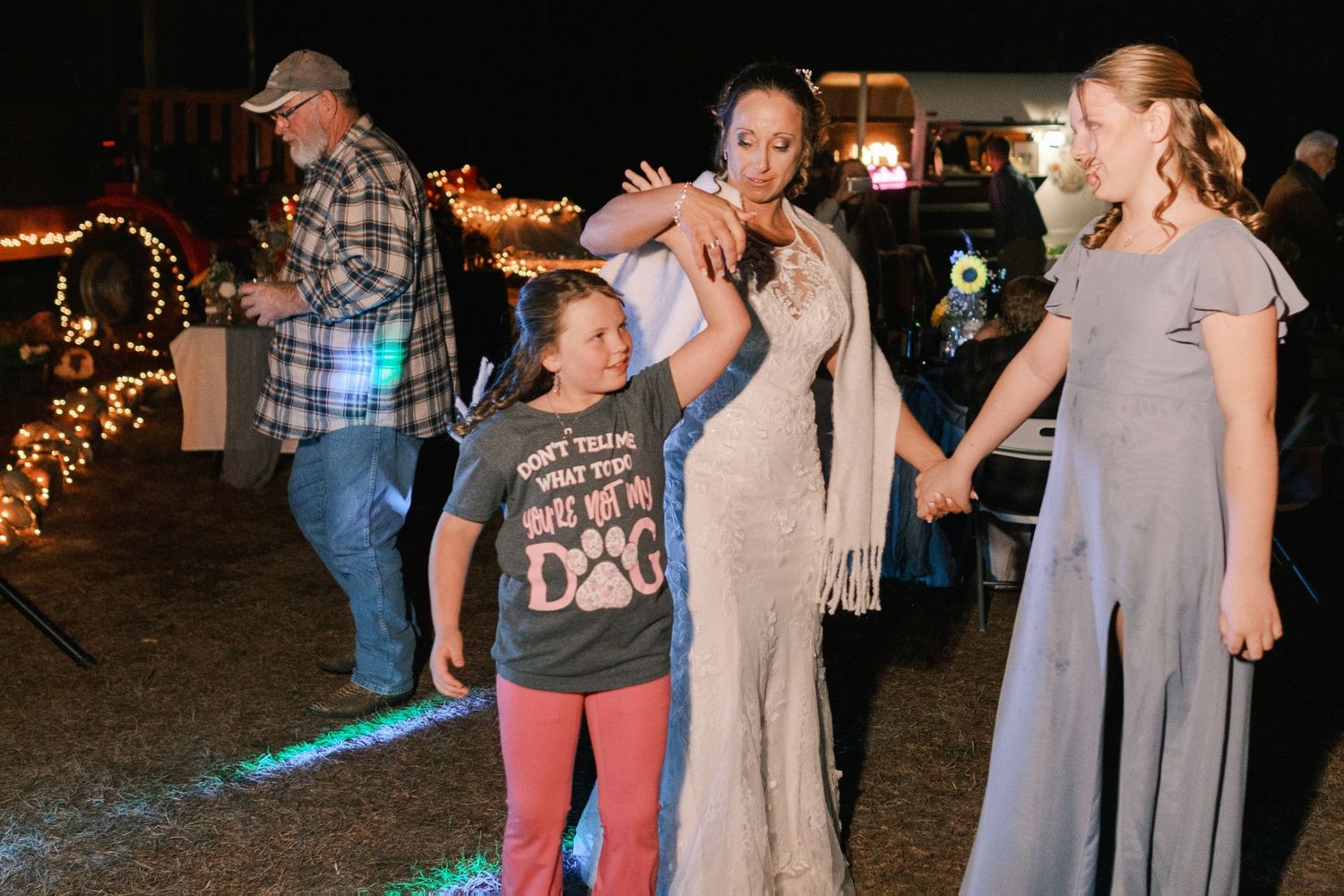 Bride in sequined gown dances with two young girls under colorful lights at an outdoor wedding reception.