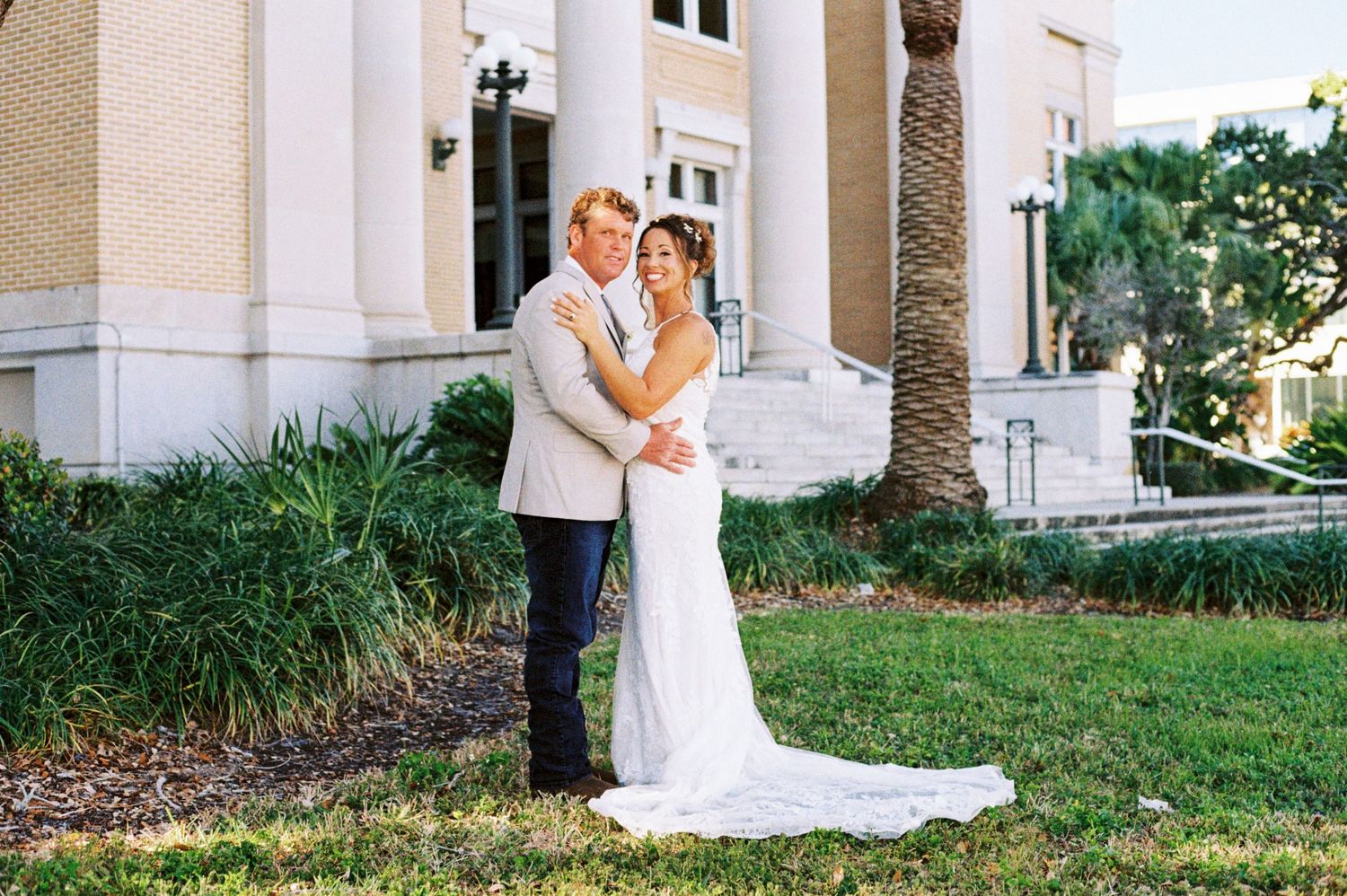 Bride and groom embrace in front of a grand building surrounded by palm trees.