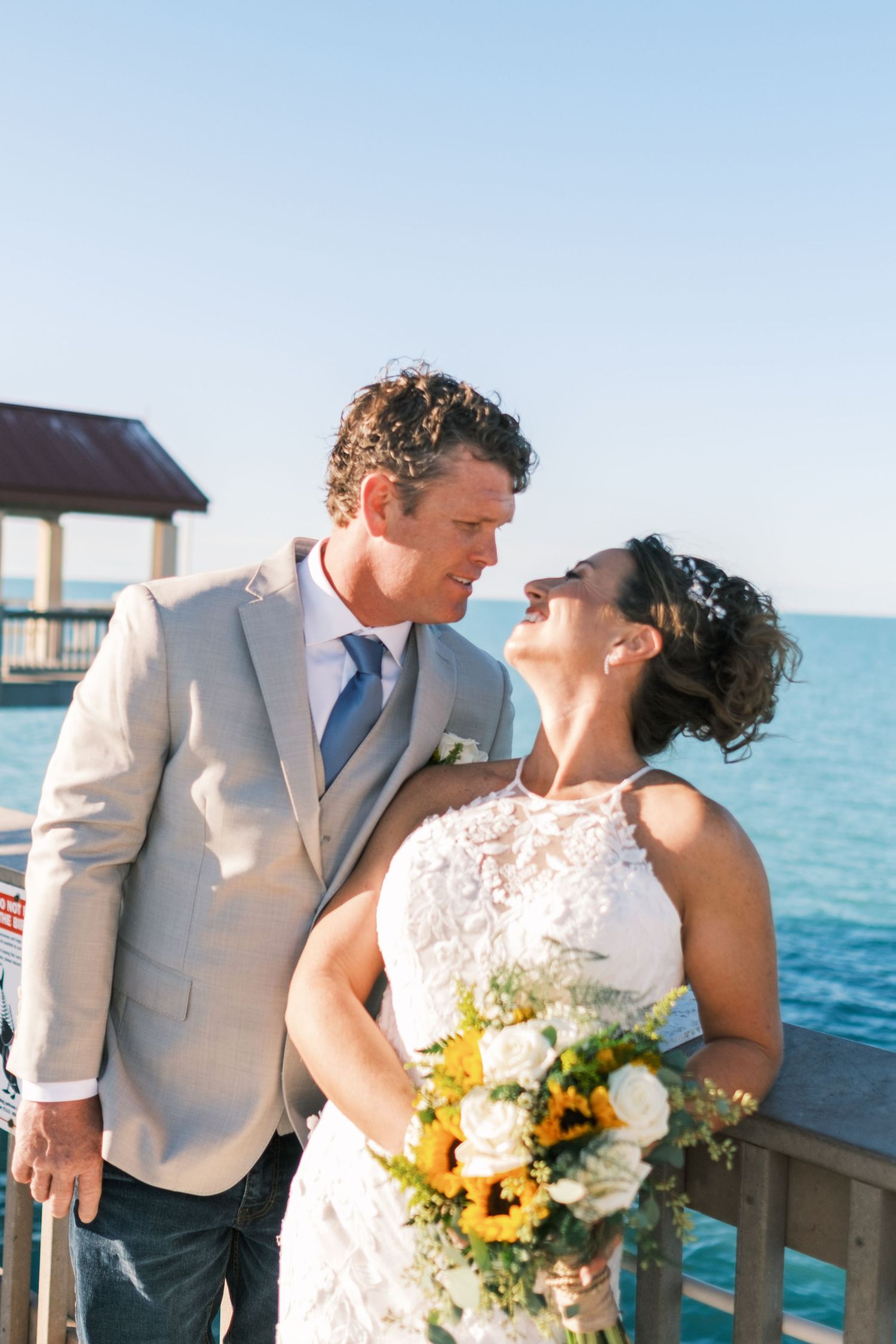 Bride and groom share romantic moment on waterfront pier, bride holding sunflower bouquet.