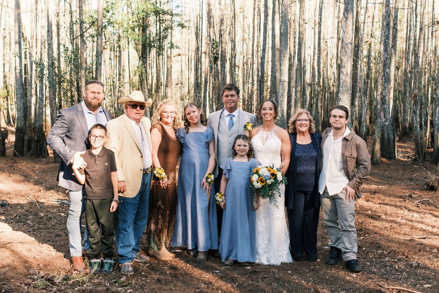 Wedding party group photo in a pine forest with bride, groom, bridesmaids in blue dresses and family.